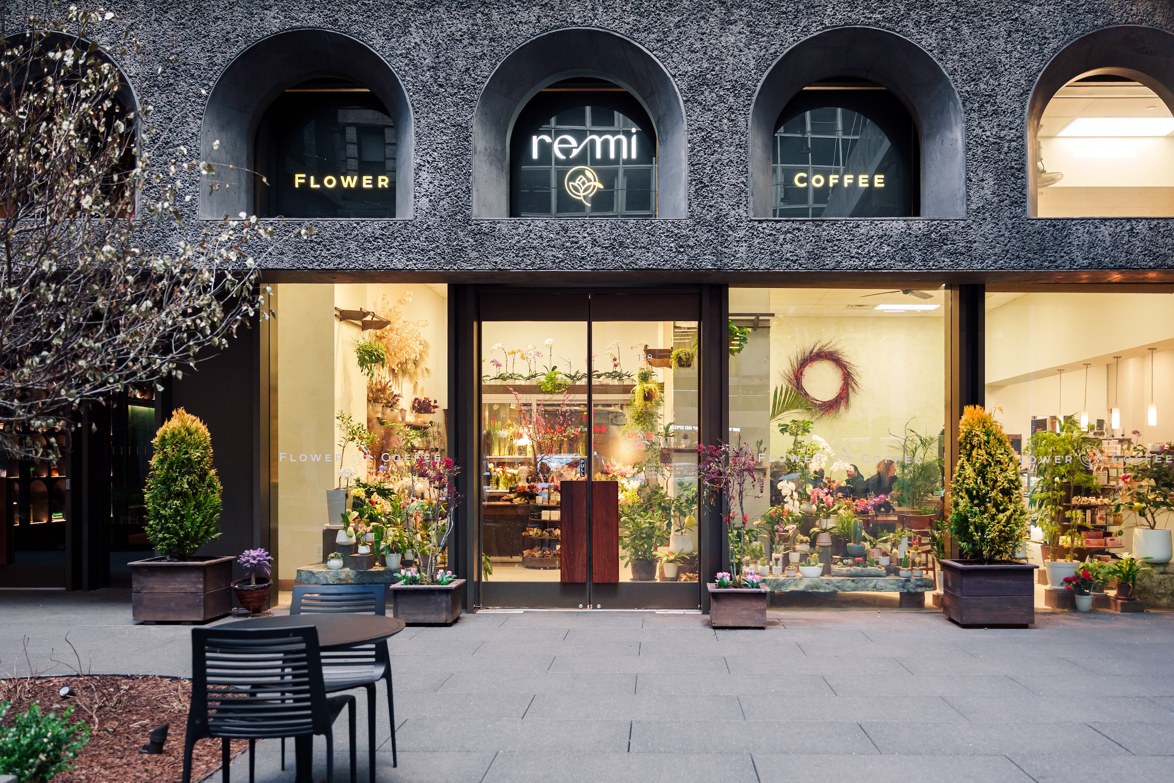 Exterior view of Remi Flower & Coffee, with large front windows showcasing vibrant floral displays and a seating area visible inside.