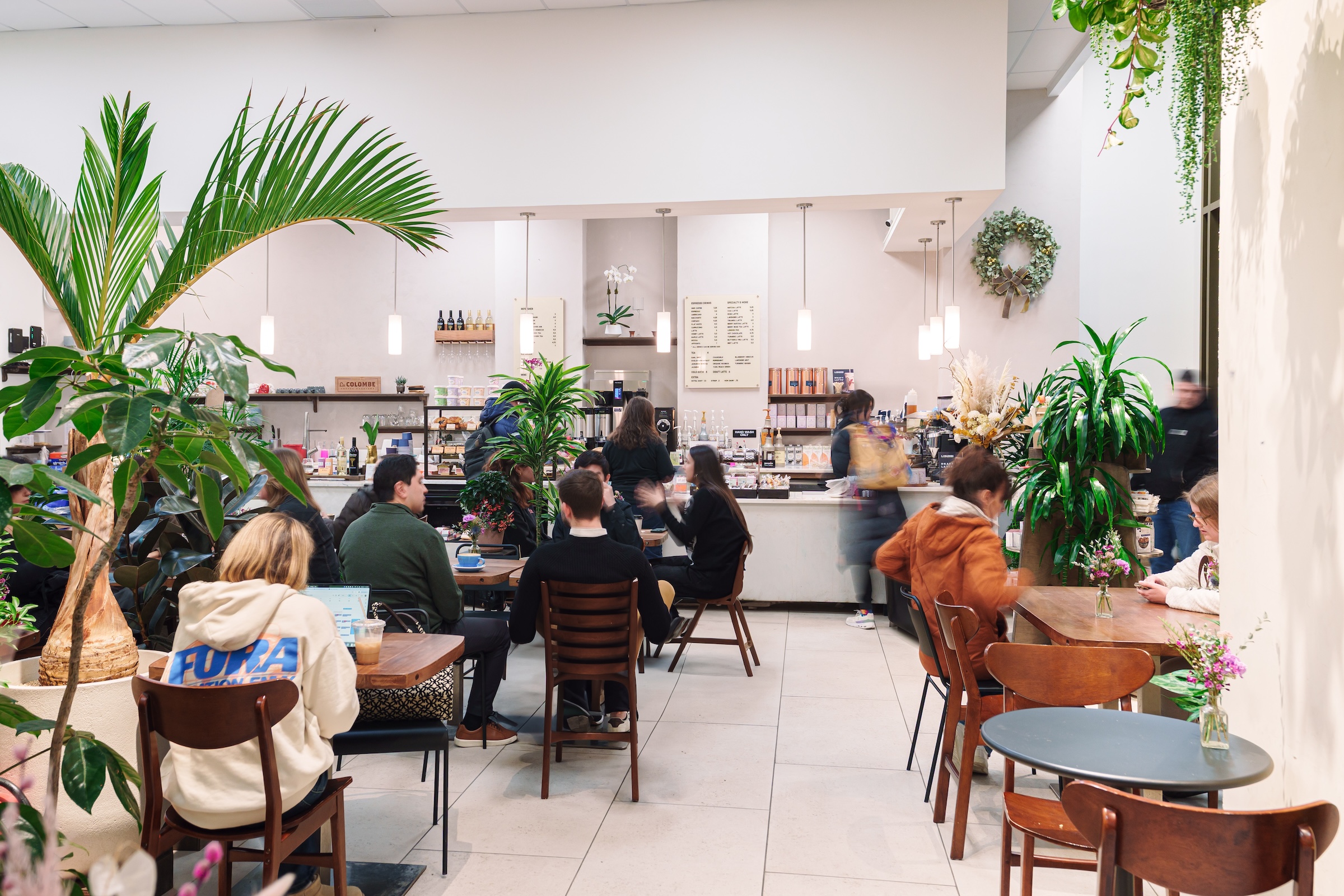 Customers sit at small wooden tables inside a bright, plant-filled café, with a counter in the background displaying drinks, pastries, and a menu board.