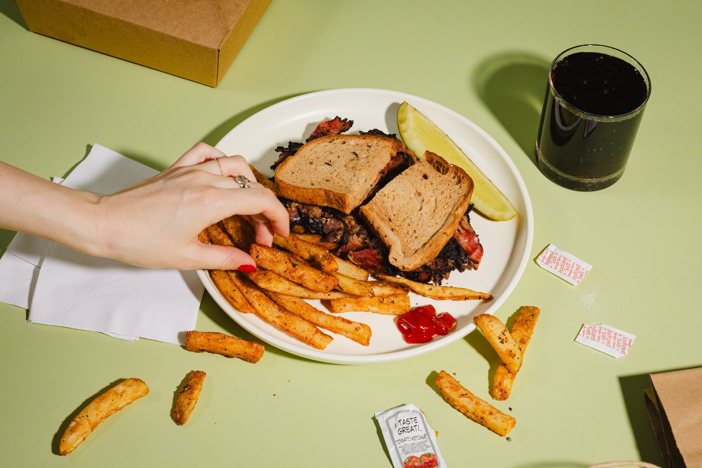 A plate of a pastrami or brisket sandwich on rye with seasoned fries, a pickle spear, and ketchup, with a hand reaching for a fry. A glass of soda, packets of ketchup, and scattered fries sit on a pale green table.