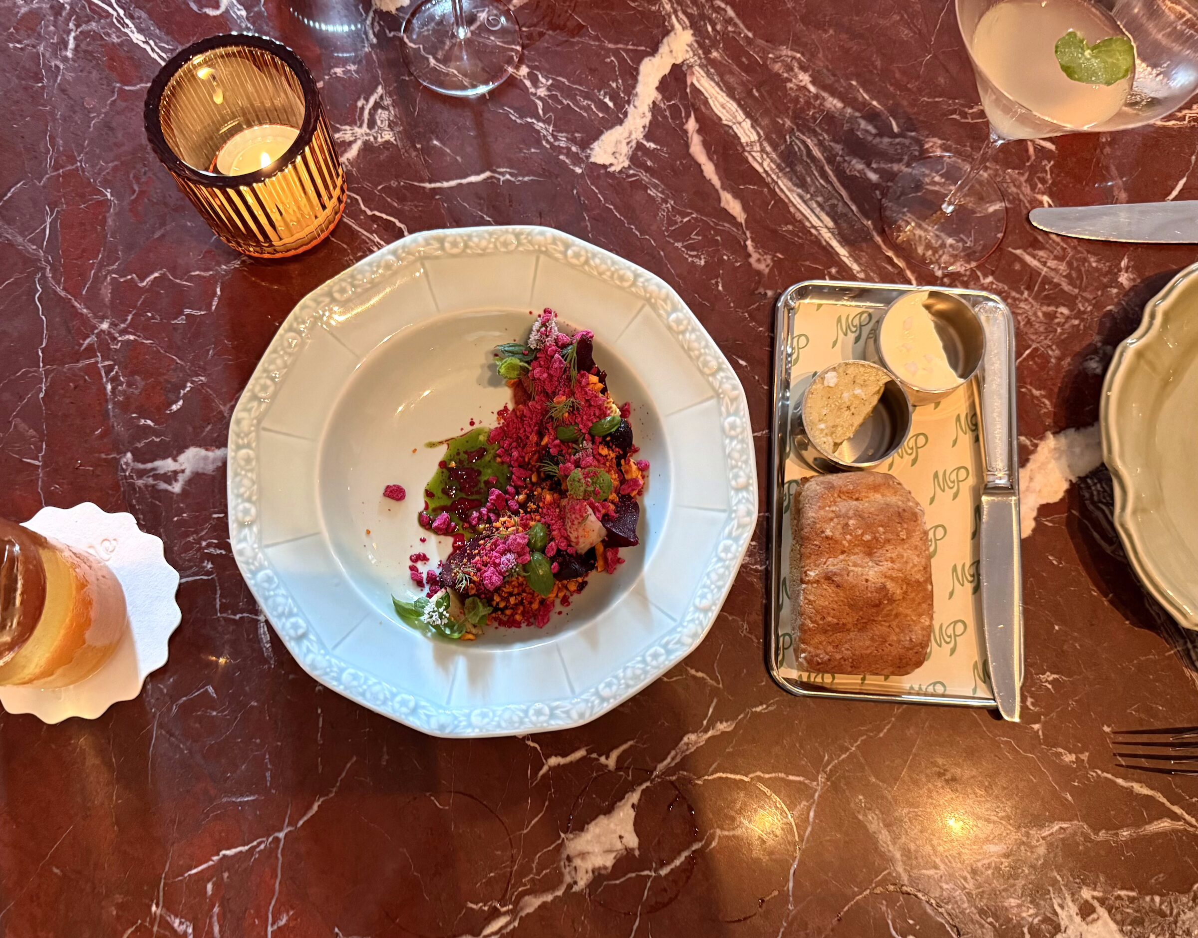 A tray with a small loaf of bread and a knife next to a plate with a colorful beet salad, all atop a red marble table.