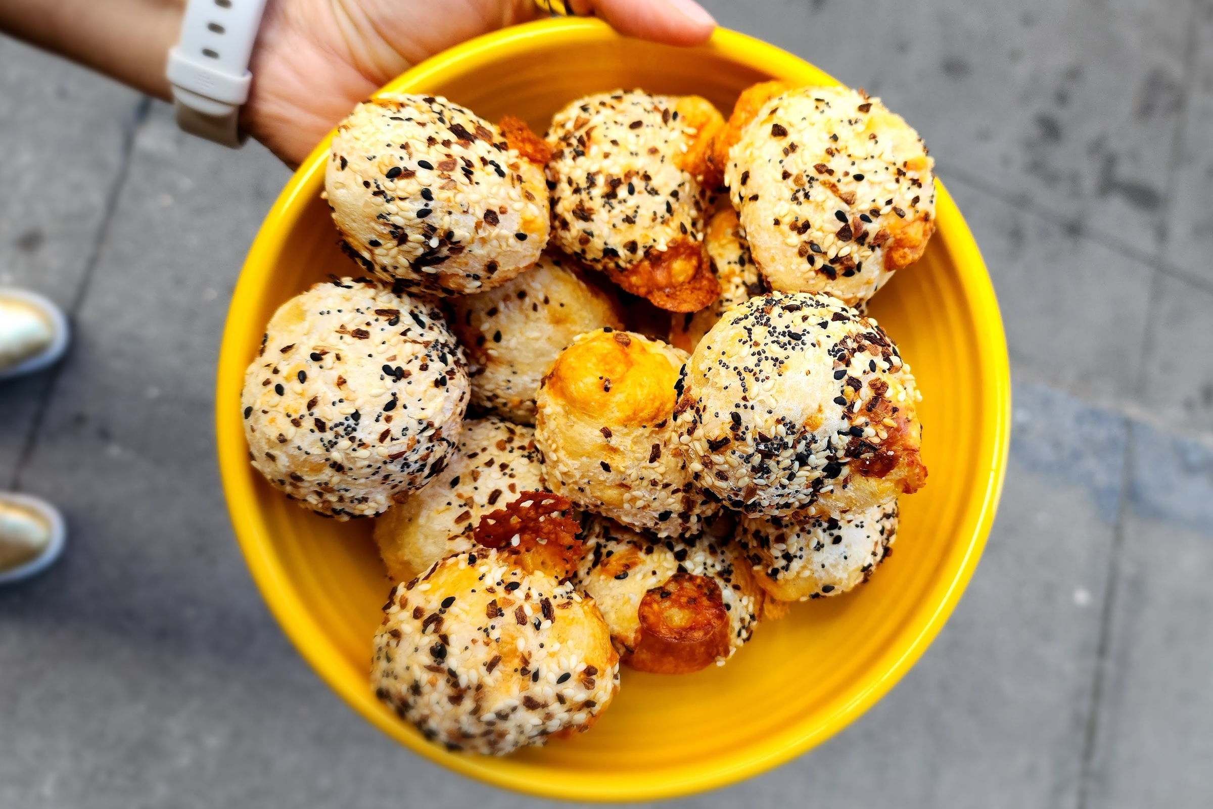 A hand holding a bright yellow bowl filled with golden-brown, everything bagel-seasoned cheese puffs, with a sidewalk background visible below.