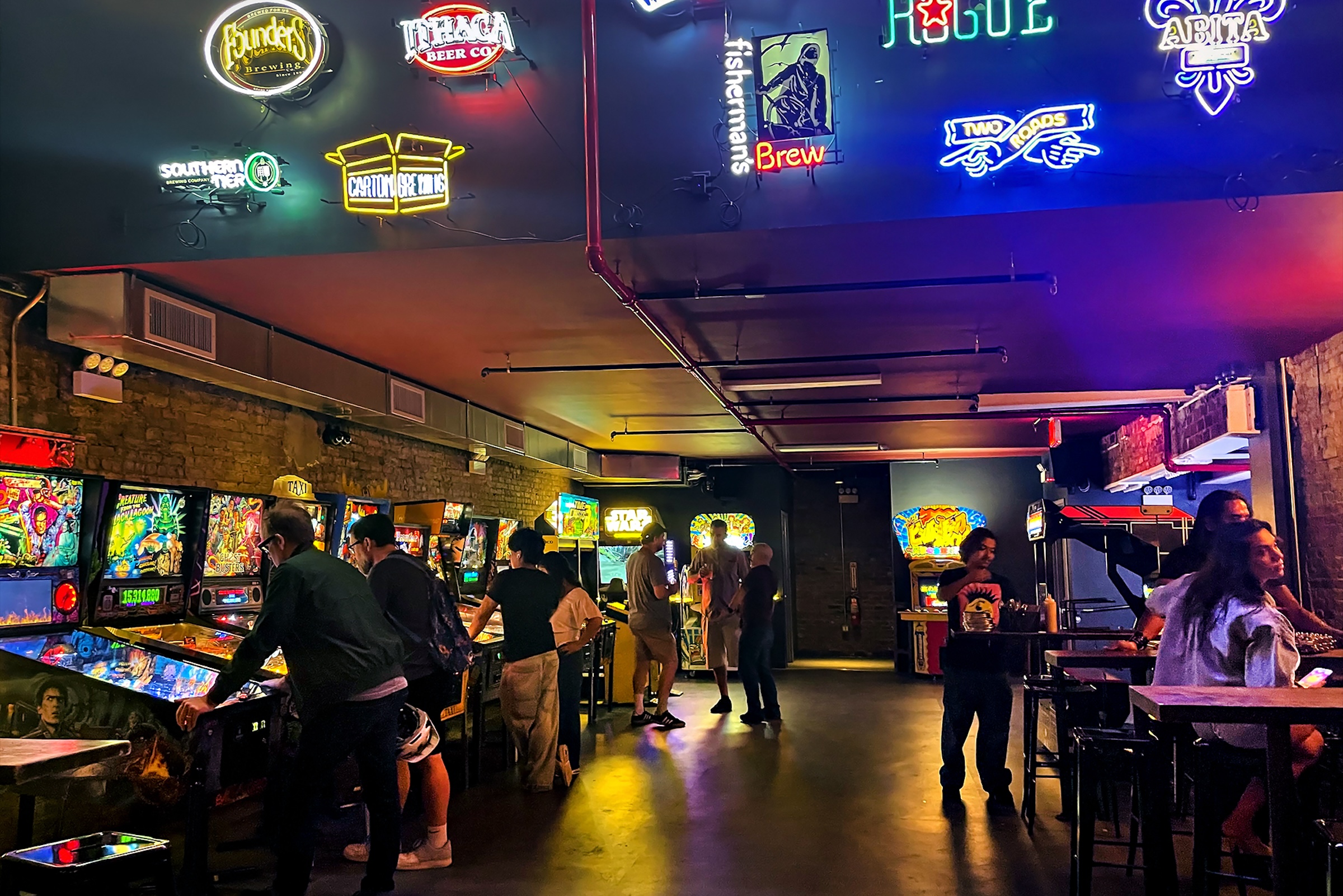 People playing pinball and arcade games in a warmly lit bar arcade, with neon beer signs hanging on the ceiling and walls.