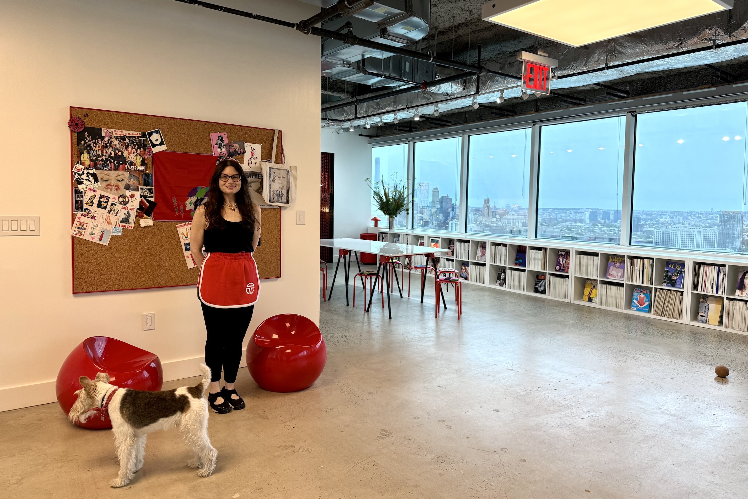 A woman in a black shirt and red skirt stands in front of a corkboard with a dog, a wall of windows and bookshelves full of magazines in the background.