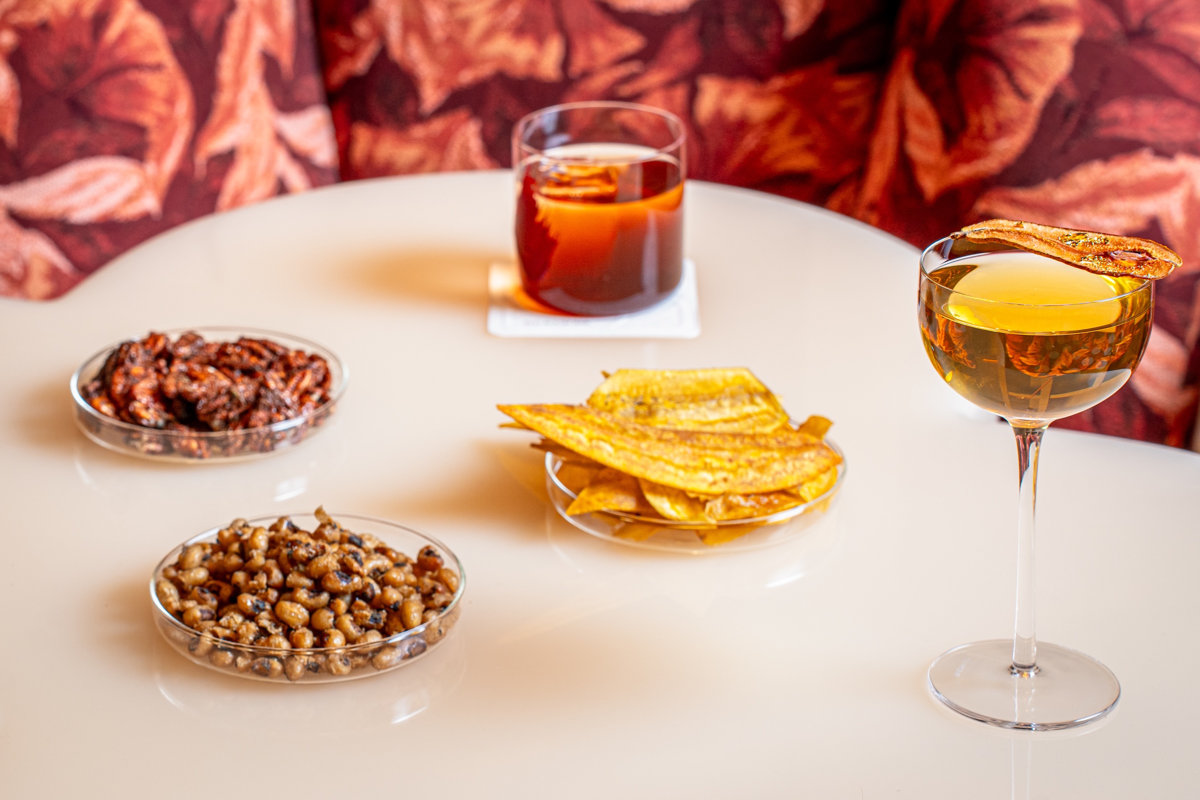 A tabletop spread with cocktails and snacks, featuring a coupe glass with a golden drink garnished with a banana chip, a tumbler with a dark cocktail on a coaster, and small dishes of plantain chips, candied nuts, and roasted legumes.