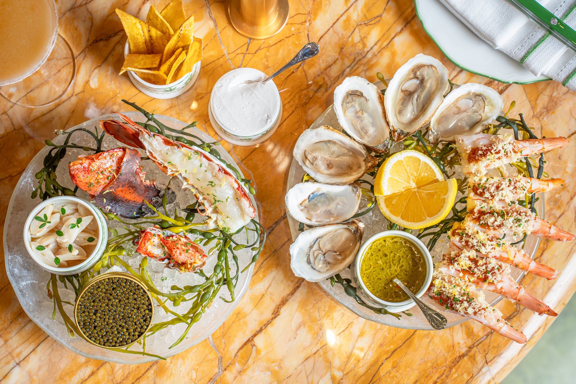 A seafood platter served on ice, featuring oysters with lemon wedges, shrimp topped with garnish, lobster pieces, caviar, and pickled vegetables, accompanied by dipping sauces and plantain chips on a marble table.