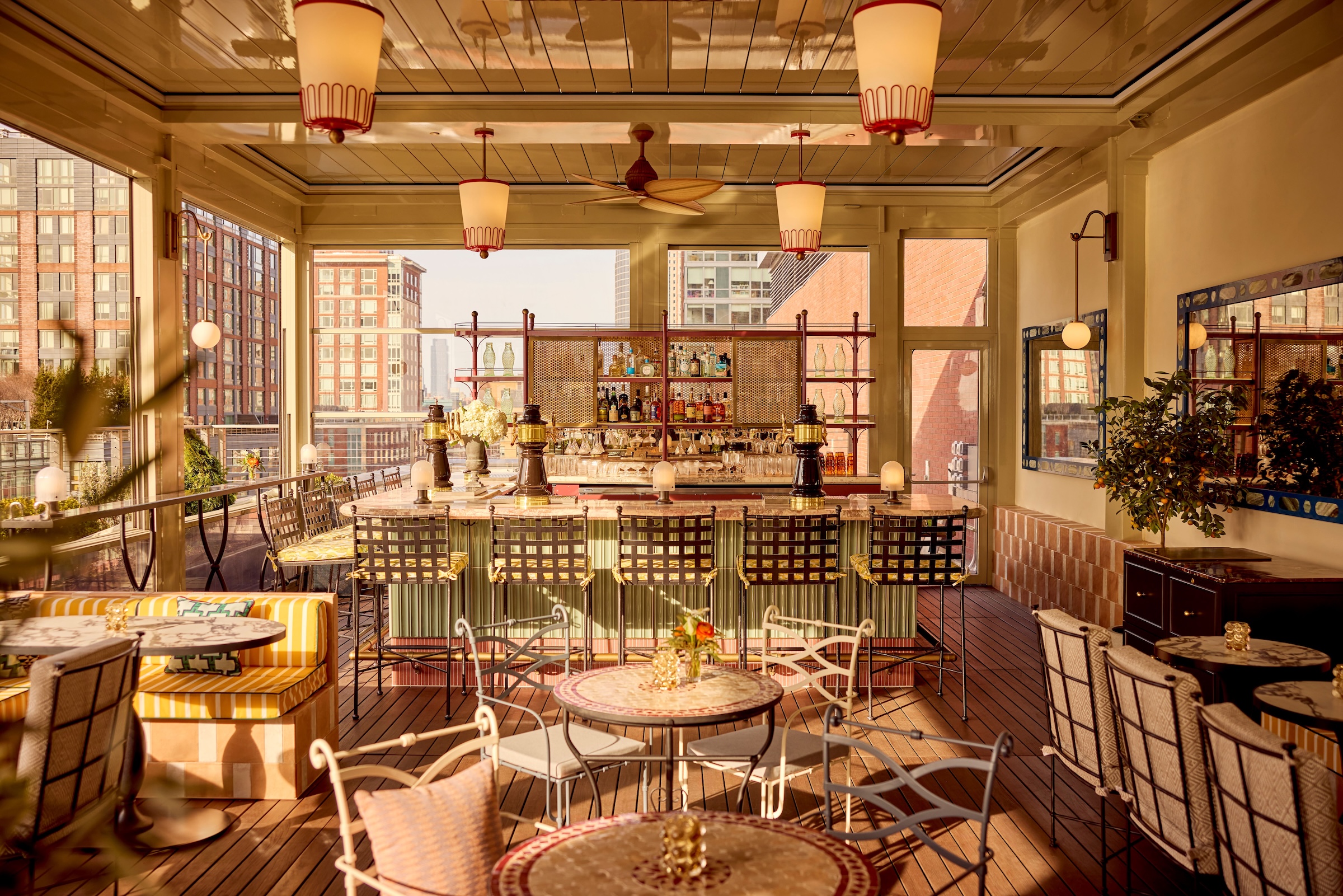 A stylish rooftop bar with striped yellow-and-white banquettes, marble-topped tables, and cushioned chairs. The bar is framed with wicker accents and bottles on display, while large windows offer views of city buildings in the background.