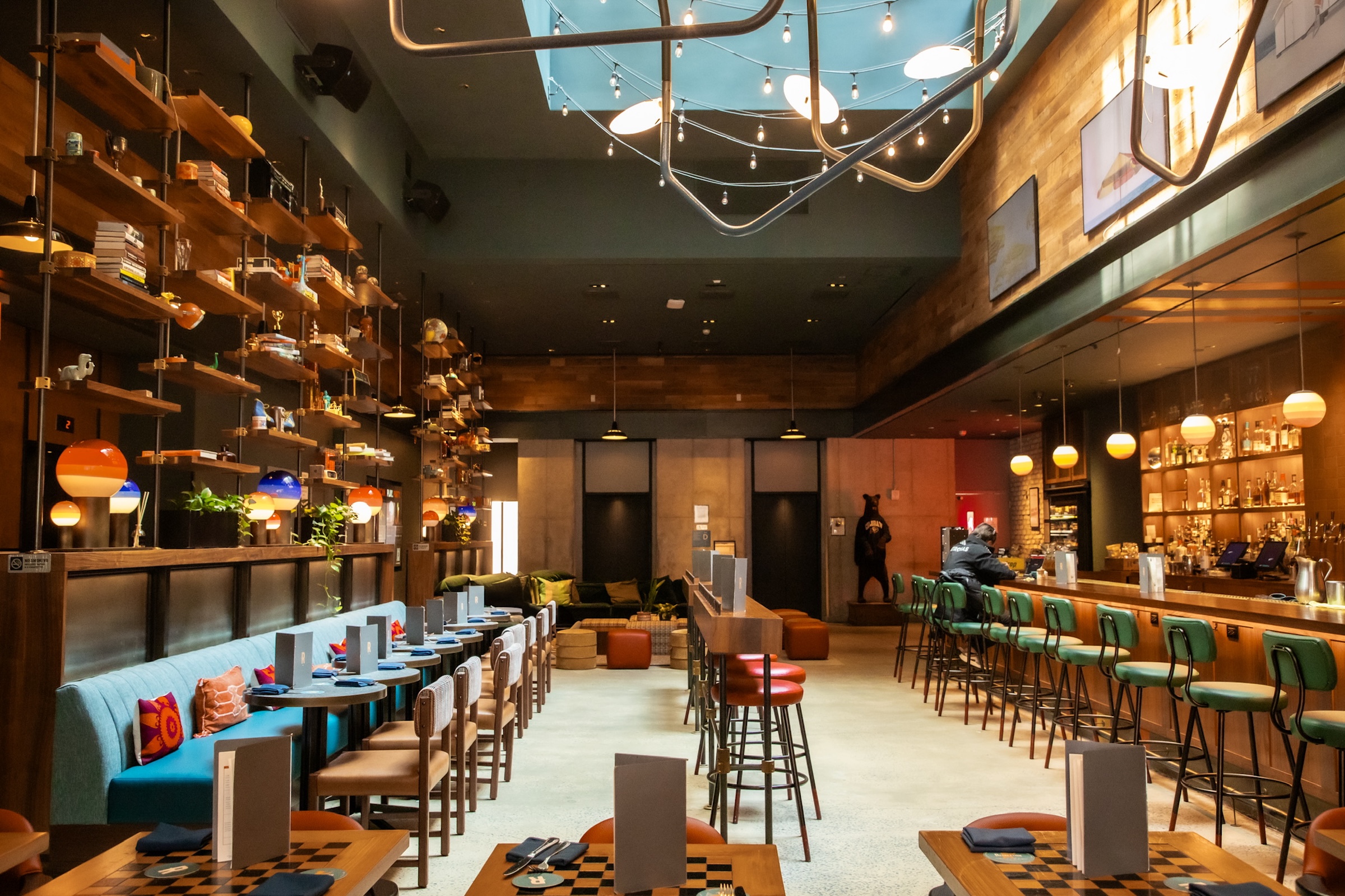A large hotel bar with a row of blue tables and banquets on one side and a stylish wood bar with green chairs on the other.