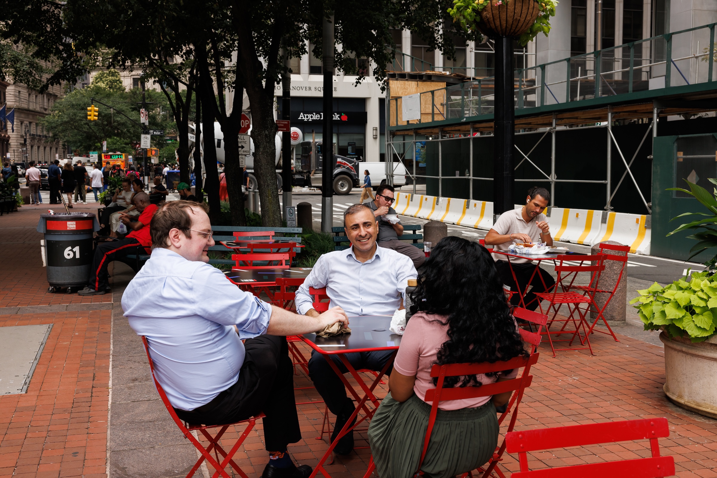 Three people sit at a red table on the brick-paved plaza at Old Slip Park in Lower Manhattan, chatting over lunch. Other people sit at nearby tables and benches, with trees, planters, and city buildings in the background.