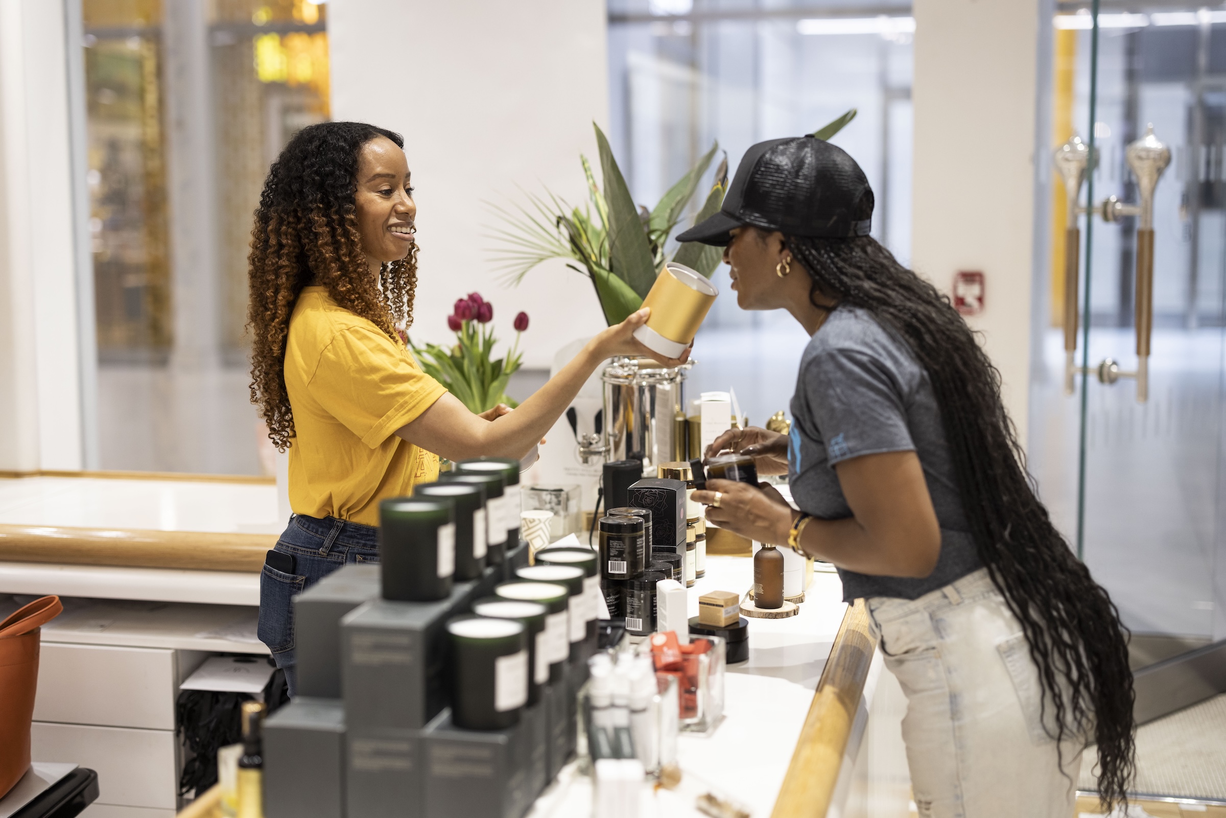 Woman in shop shows customer wares with product in the foreground and a window in the background.
