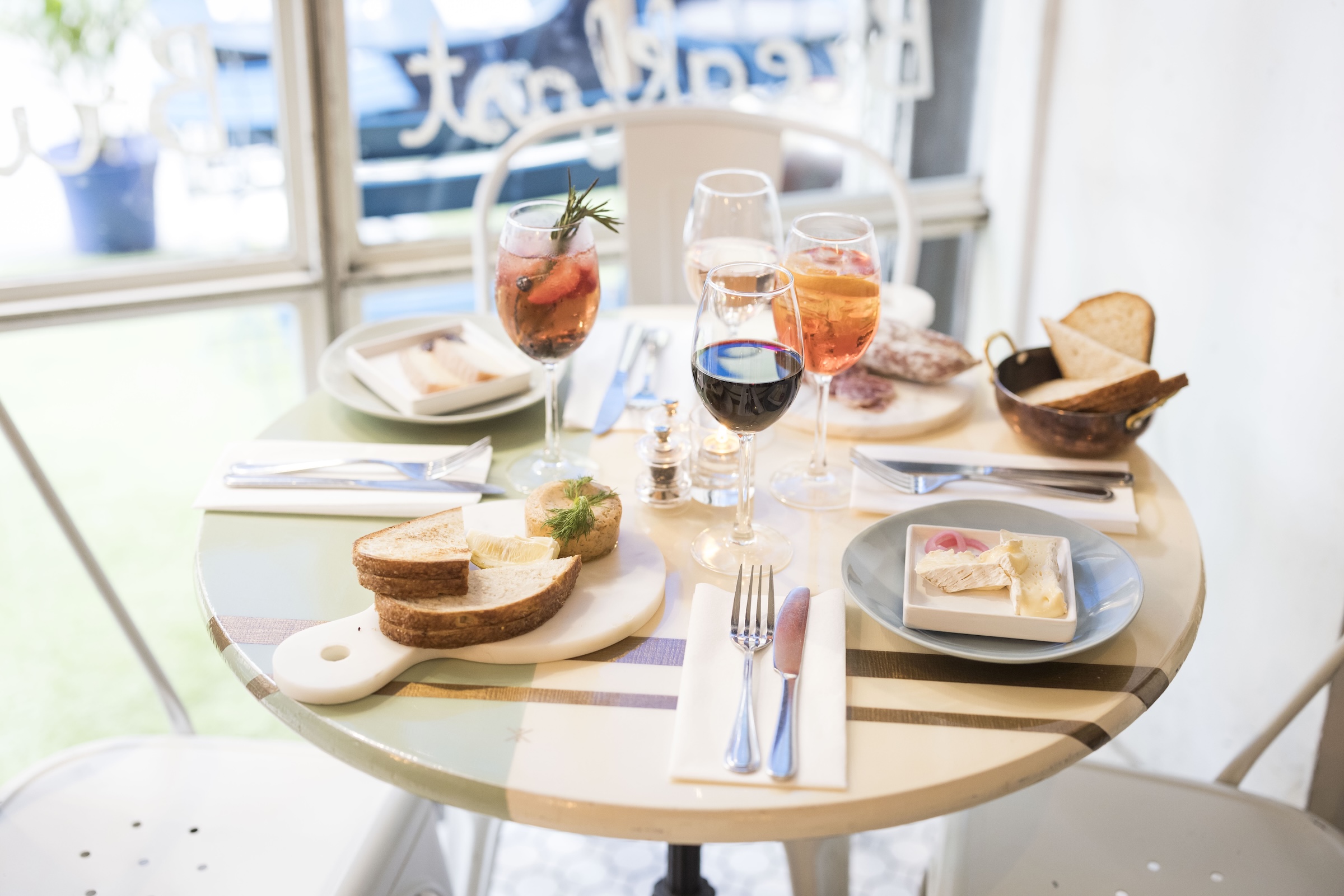 Set table at sunlit restaurant with bread, butter, cheese, salami, wine and aperol spritzes. 