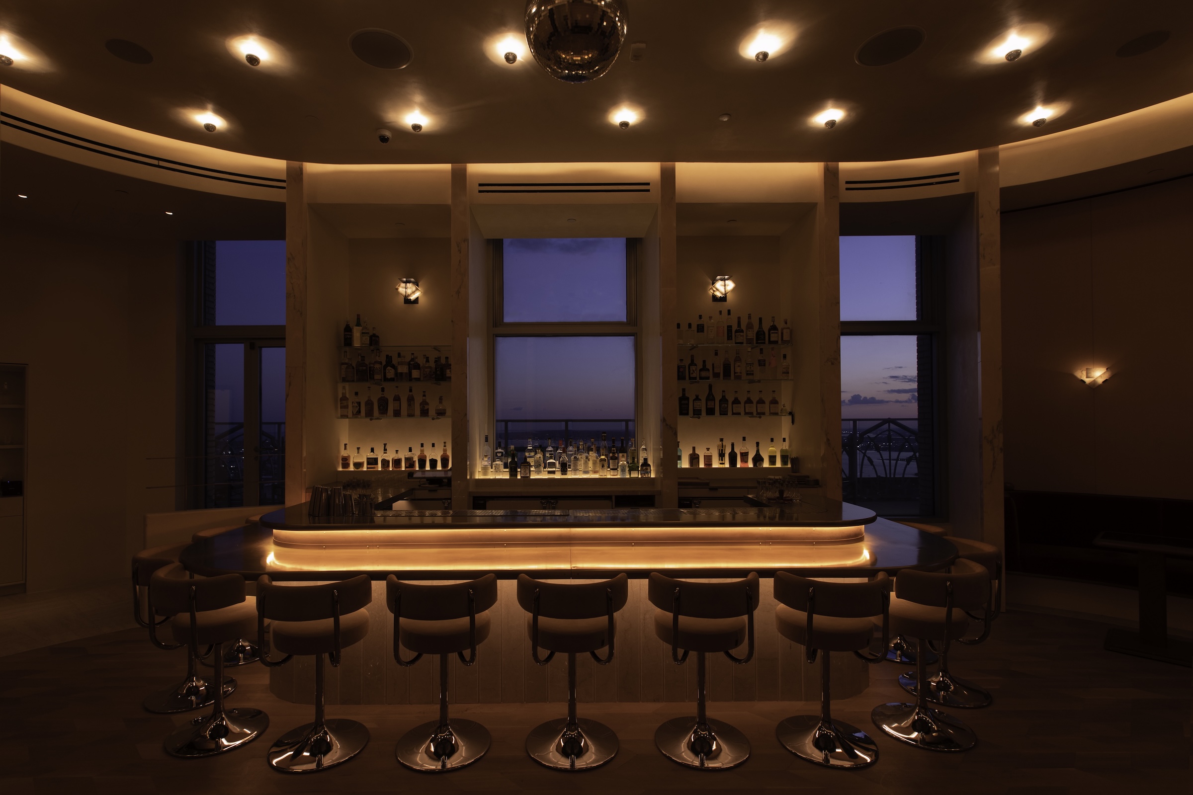 Softly backlit bar with bar stools and view of sunset in the background.