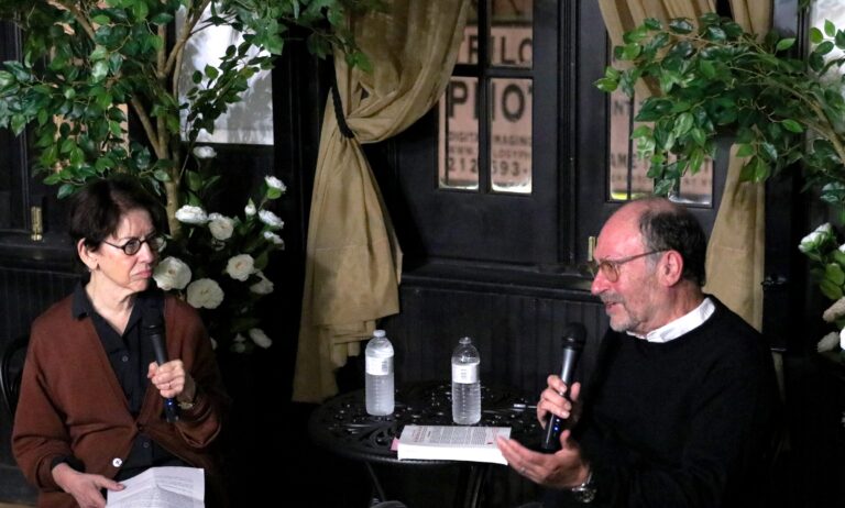 A woman and a man hold microphones while sitting and talking to each other in front of a black table with water bottles on it, with greenery and curtains in the background.