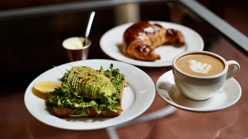 Avocado toast, croissant and cup of coffee with latte art sit on table.