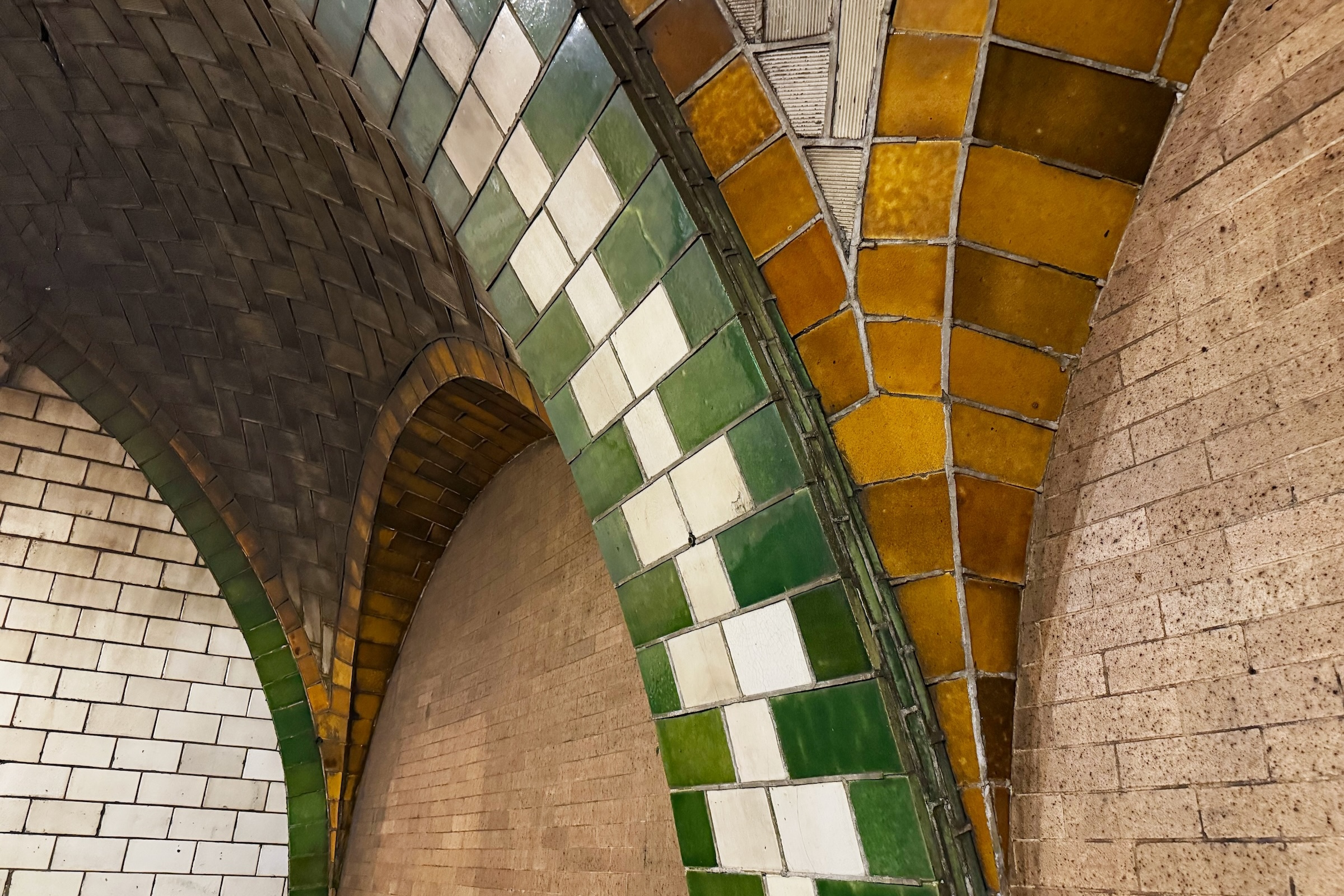 Close-up view of green and yellow tiling on arch of brick subway wall and ceiling.