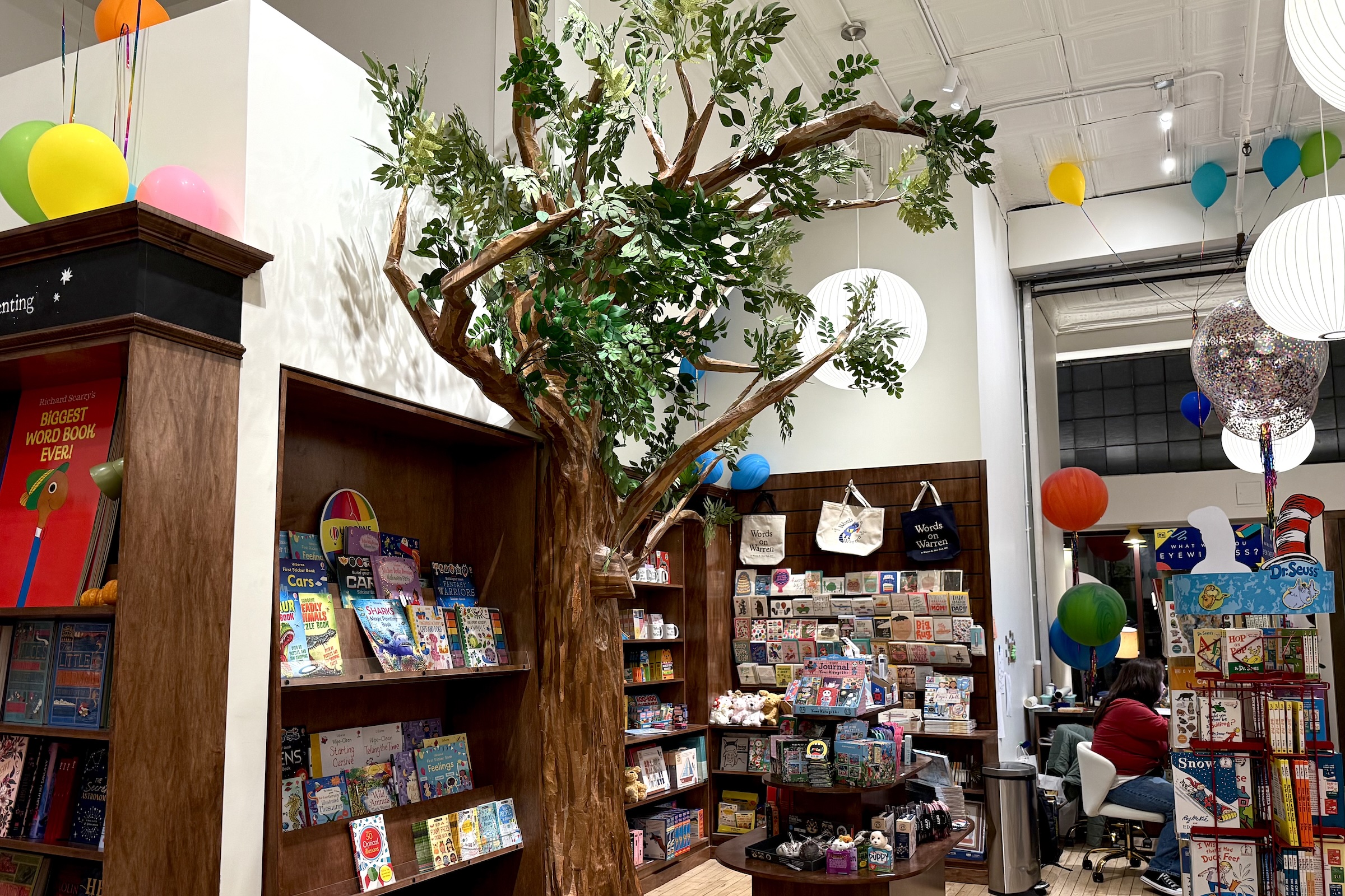 A brightly lit children’s bookstore interior featuring a large artificial tree with leafy branches spreading across the ceiling. Colorful balloons float above book displays, and shelves are filled with picture books, toys, and gifts. A staff member sits at a desk in the back, and festive hanging lanterns add to the playful, whimsical atmosphere.