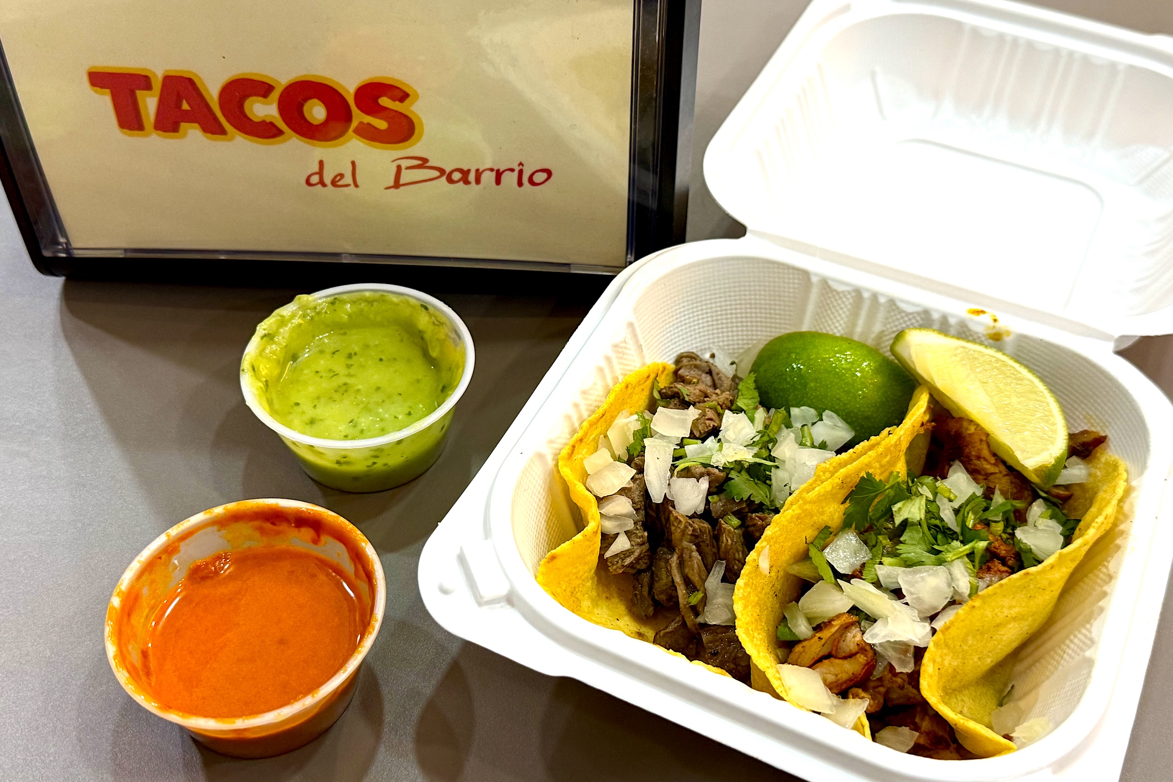A carton with two tacos next to small containers with green and red sauce. A sign reading Tacos del Barrio sits next to them.