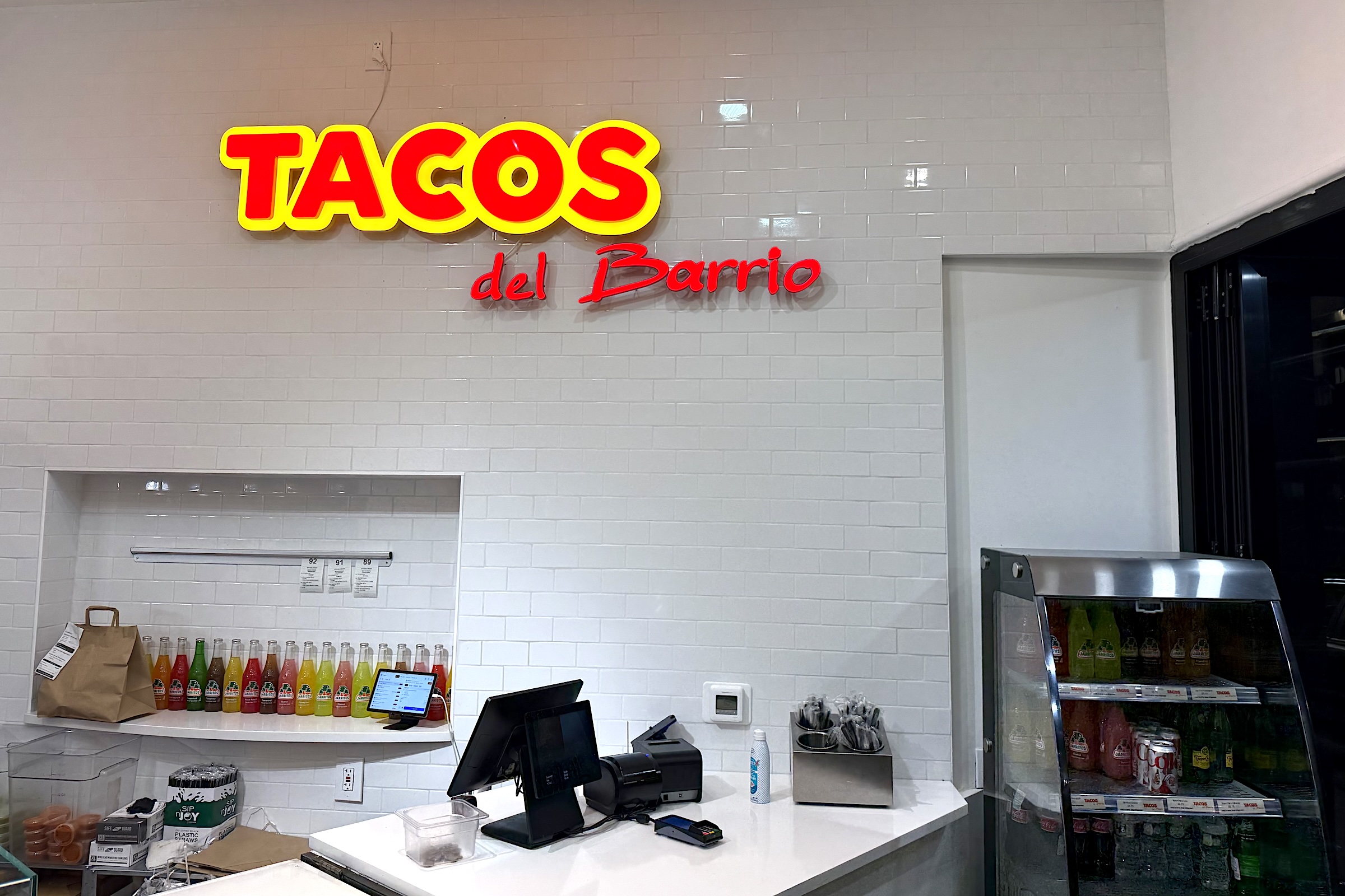A brightly lit sign reading "Tacos del Barrio" hands on a white brick wall above a shelf with colorful drinks and a register.