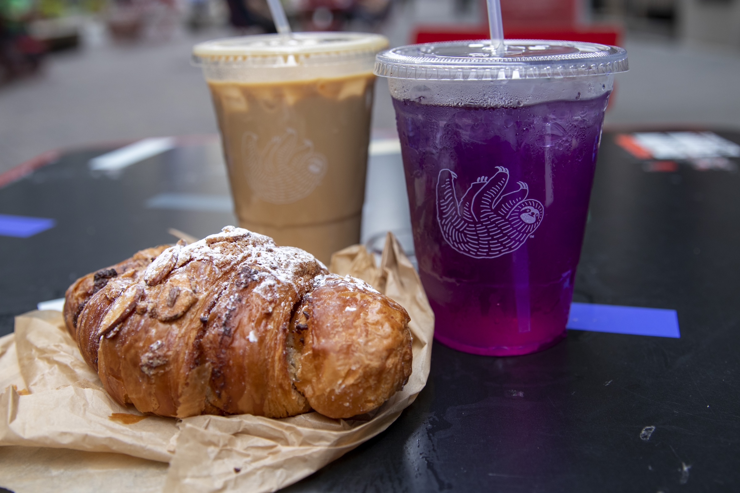 A coffee latte and purple ube drink sit next to each other on black table alongside almond croissant.