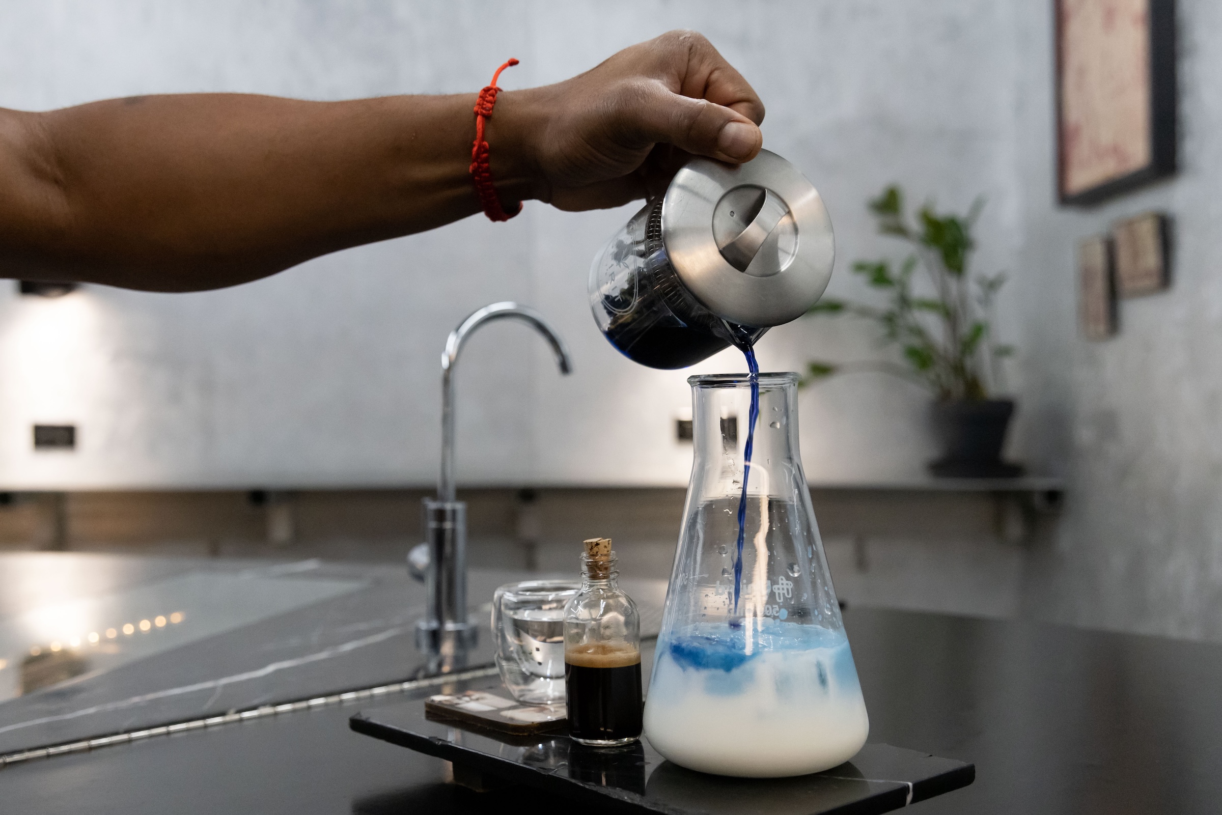 A man pours blue liquid into a beaker full of milk with a shot of coffee and a small glass of water.