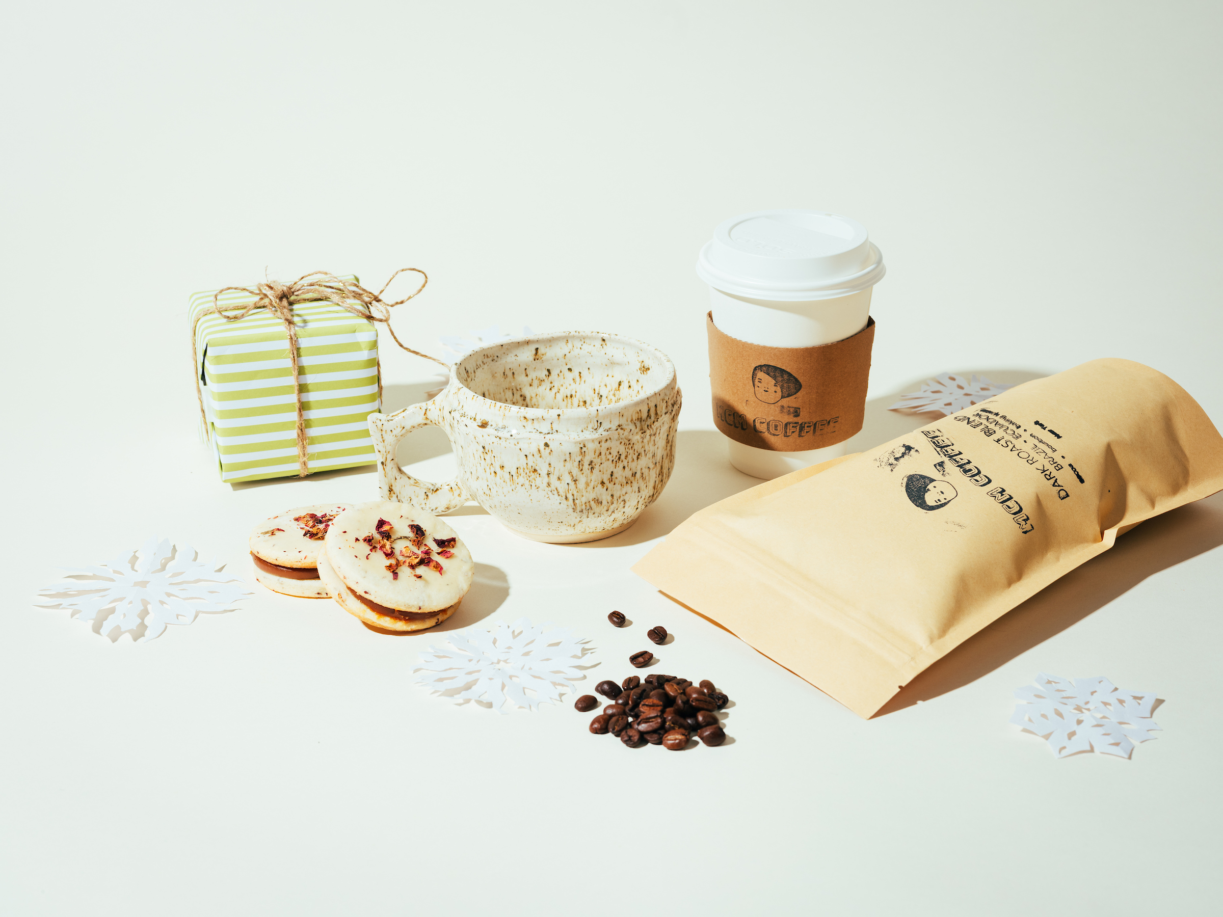 Coffee bags, cups, mugs, beans and snacks on off-white background with paper snowflakes.
