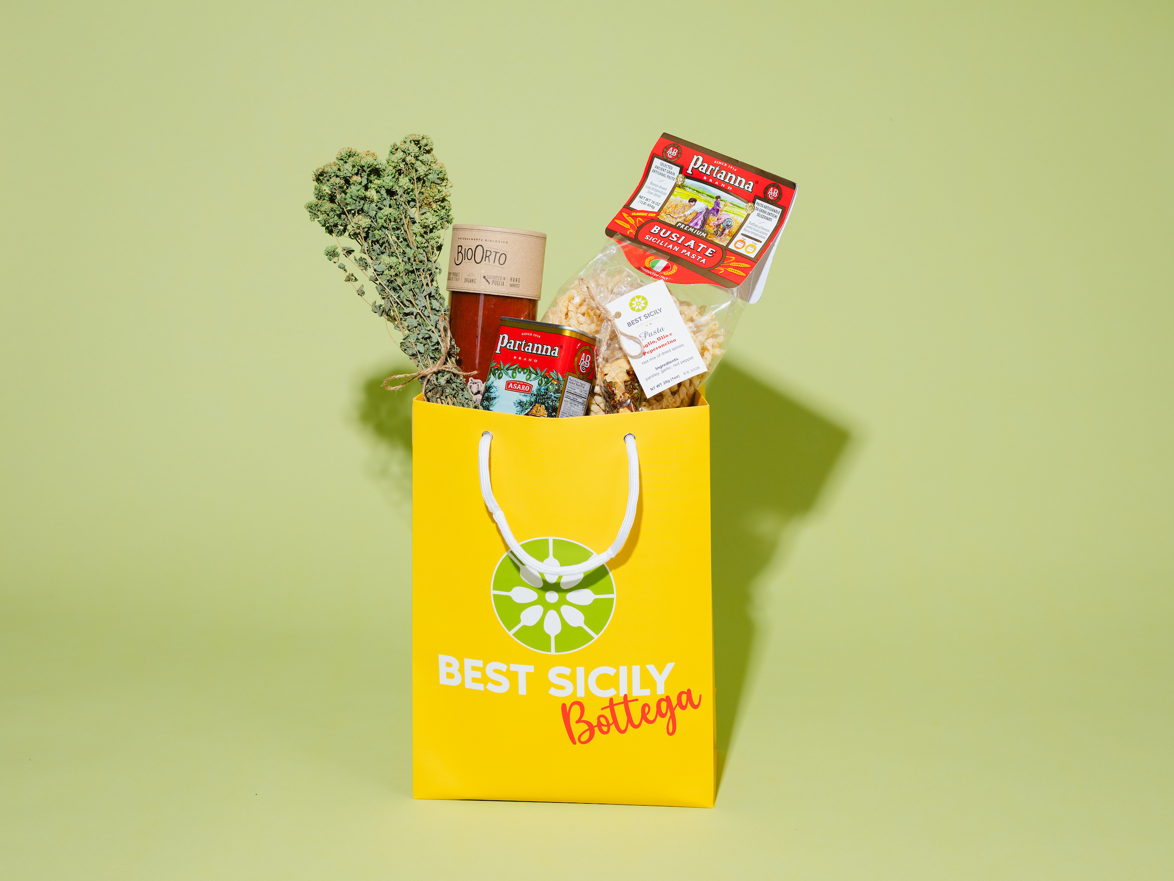 Yellow bag reading "Best Sicily Bottega" containing oregano, anchovies, chili flakes, olive oil and dried pasta against mint green background.