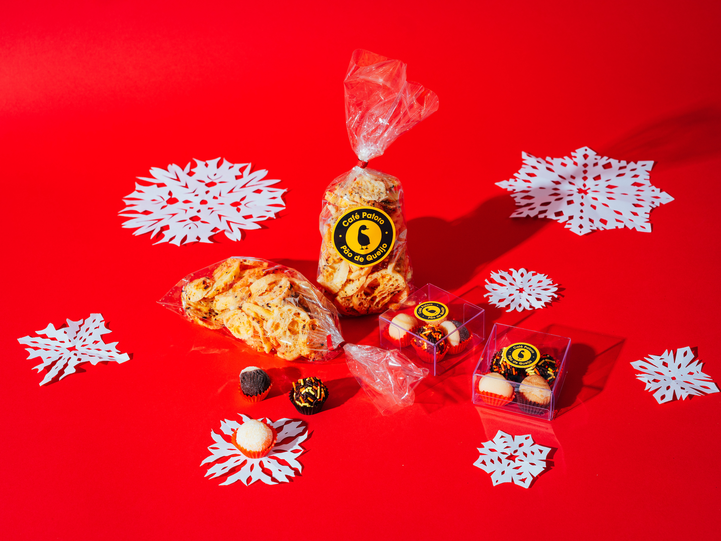 Two bags of pao de queijo reading "Cafe Patoro" and two boxes of chocolate treats against red backdrop with paper snowflakes.