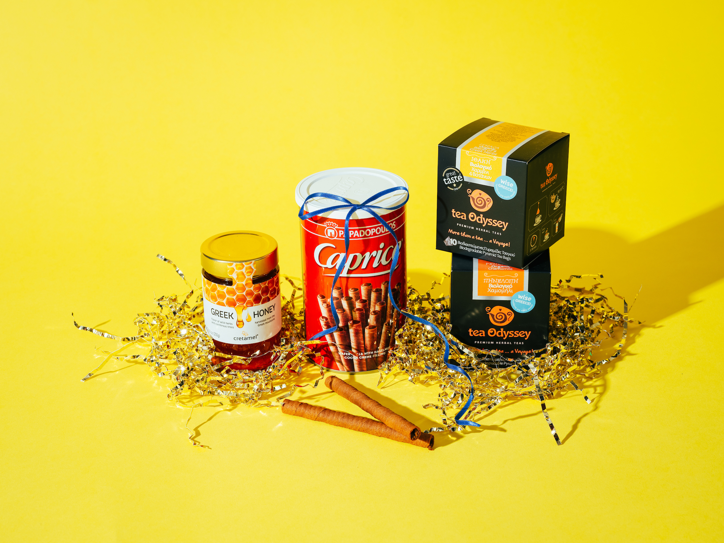 A bright yellow background displays a small arrangement of giftable food items nestled in gold and silver paper shreds. From left to right: a jar of Greek honey with a yellow lid, a tall red tin of Caprice rolled wafer cookies tied with a blue ribbon, and two stacked black boxes of Tea Odyssey herbal tea. Two cinnamon sticks rest in front of the items.