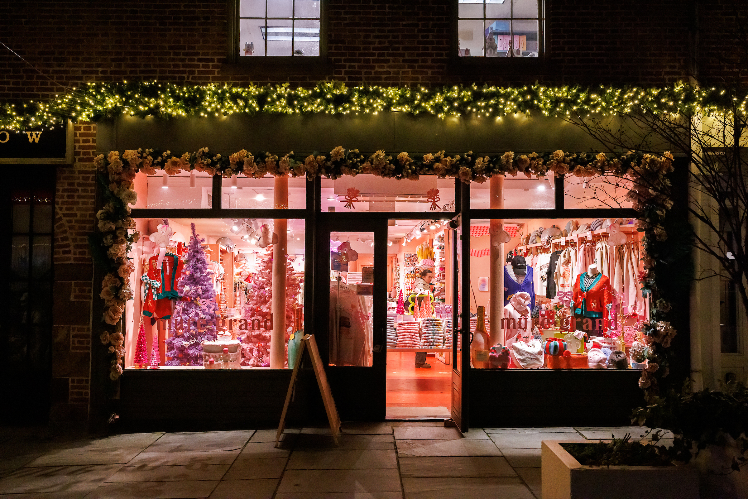 A storefront reading "Mure & Grand" is decked out in Christmas lights and wreaths, with the store's interior pink walls visible through the glass.