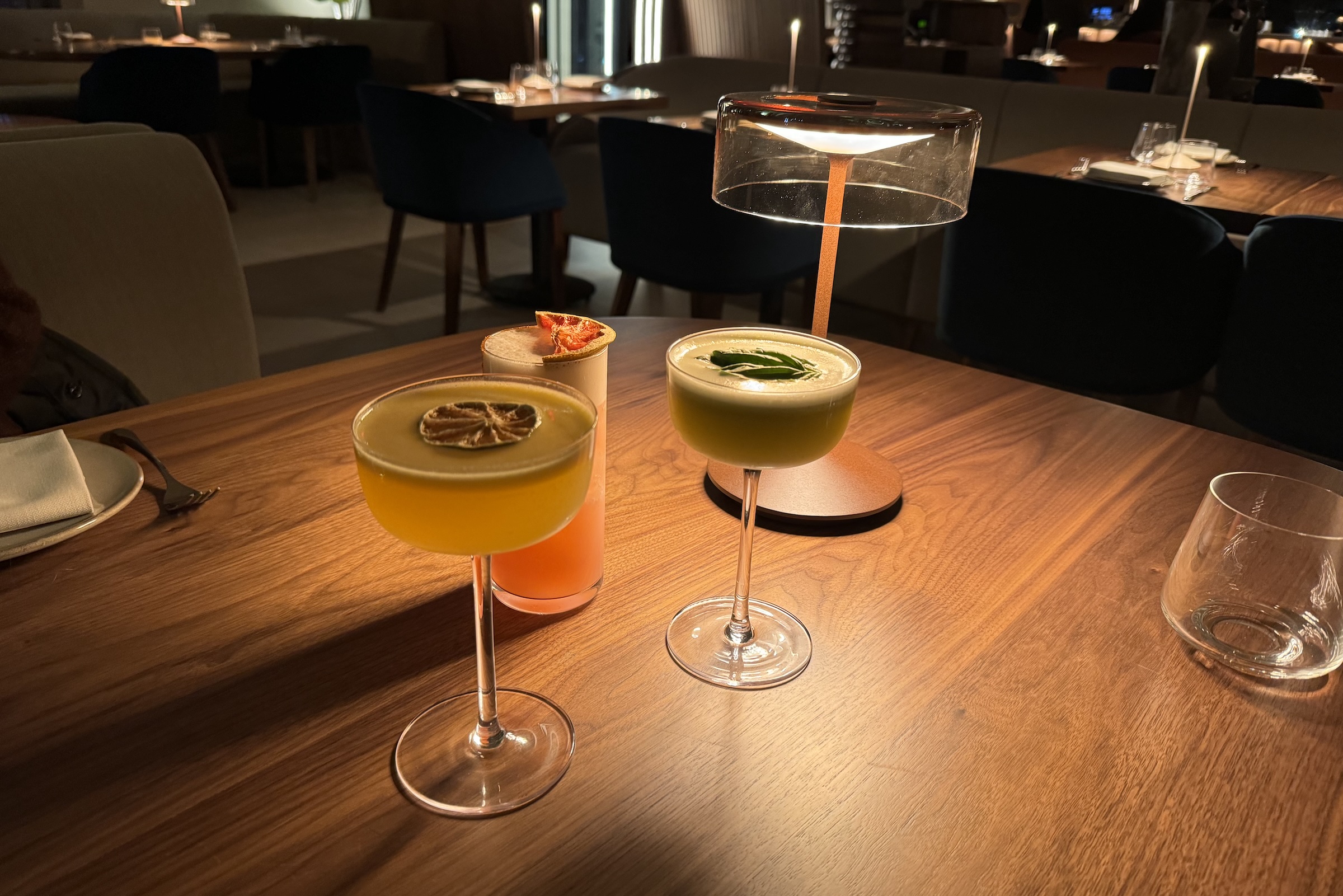 Three craft cocktails are arranged on a dark wood table in a dimly lit, upscale restaurant. One drink is a yellow cocktail in a coupe glass topped with a dried citrus wheel. Behind it is a tall pink drink garnished with a dried grapefruit slice. To the right is a pale green cocktail topped with fresh basil leaves, illuminated by a modern, minimalist table lamp with a glass shade. In the blurred background, several empty dining tables and dark blue chairs are visible under warm ambient lighting.