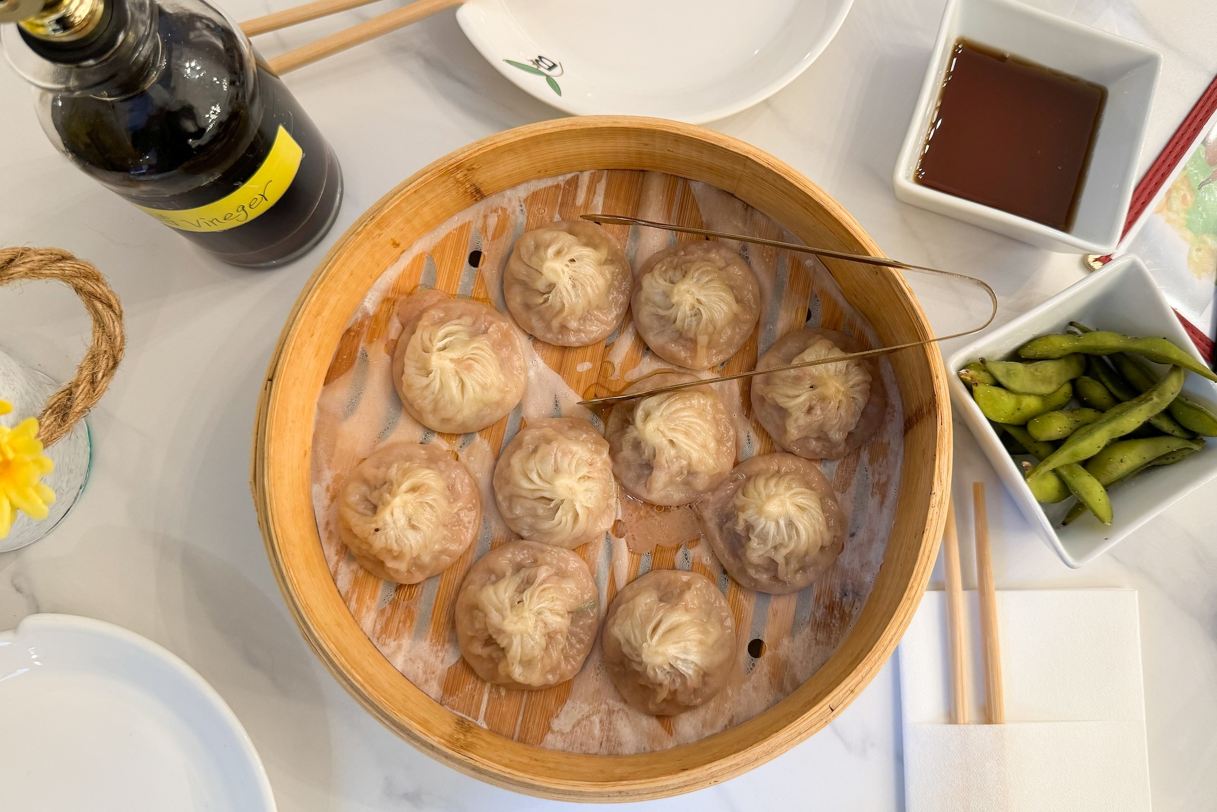 A top-down view of a light wood steamer basket containing ten Xiao Long Bao (soup dumplings) resting on parchment paper. The scene includes a side of green edamame, a small dish of soy sauce, a bottle of black vinegar, and wooden chopsticks.