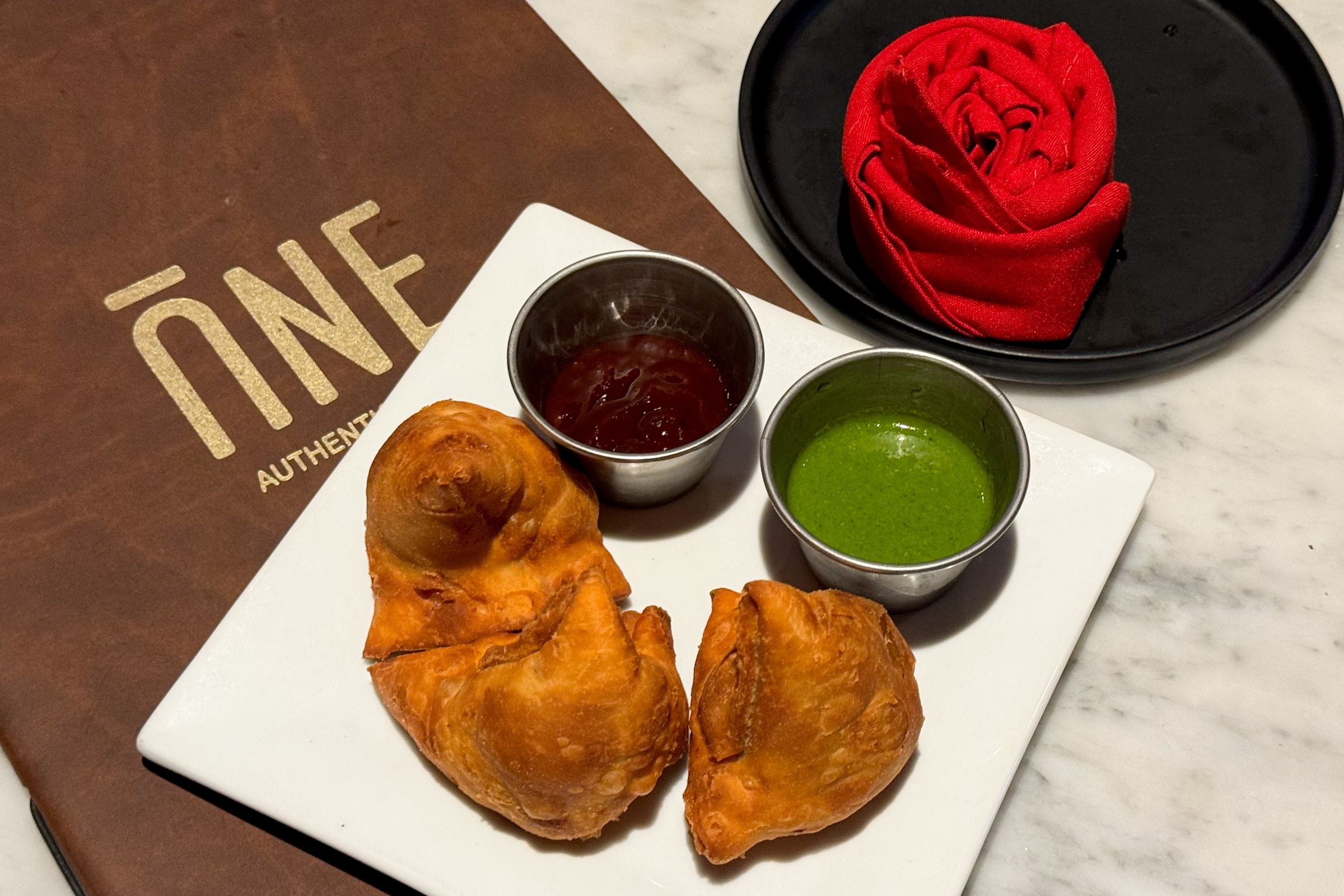 Two golden-brown, crispy samosas served on a white square plate. They are accompanied by two small metal ramekins containing dark tamarind chutney and bright green mint-cilantro chutney. In the background, there is a brown leather menu with the gold logo "ĀNĒ" and a red cloth napkin folded into a rose shape.