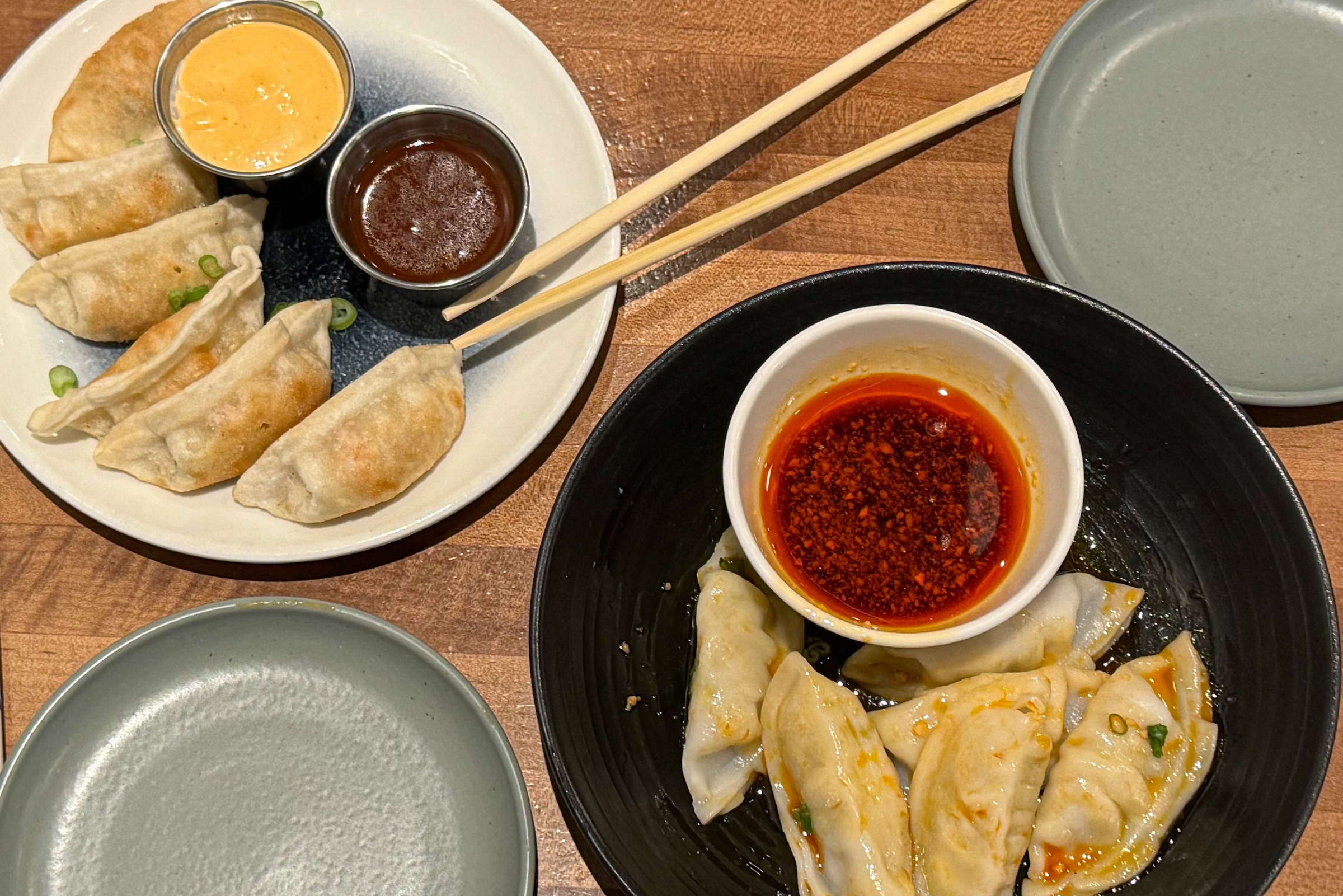 A high-angle shot of two plates of dumplings on a wooden table. The plate on the left holds pan-fried gyoza with two dipping sauces. The black bowl on the right contains steamed dumplings swimming in a spicy red chili oil sauce.