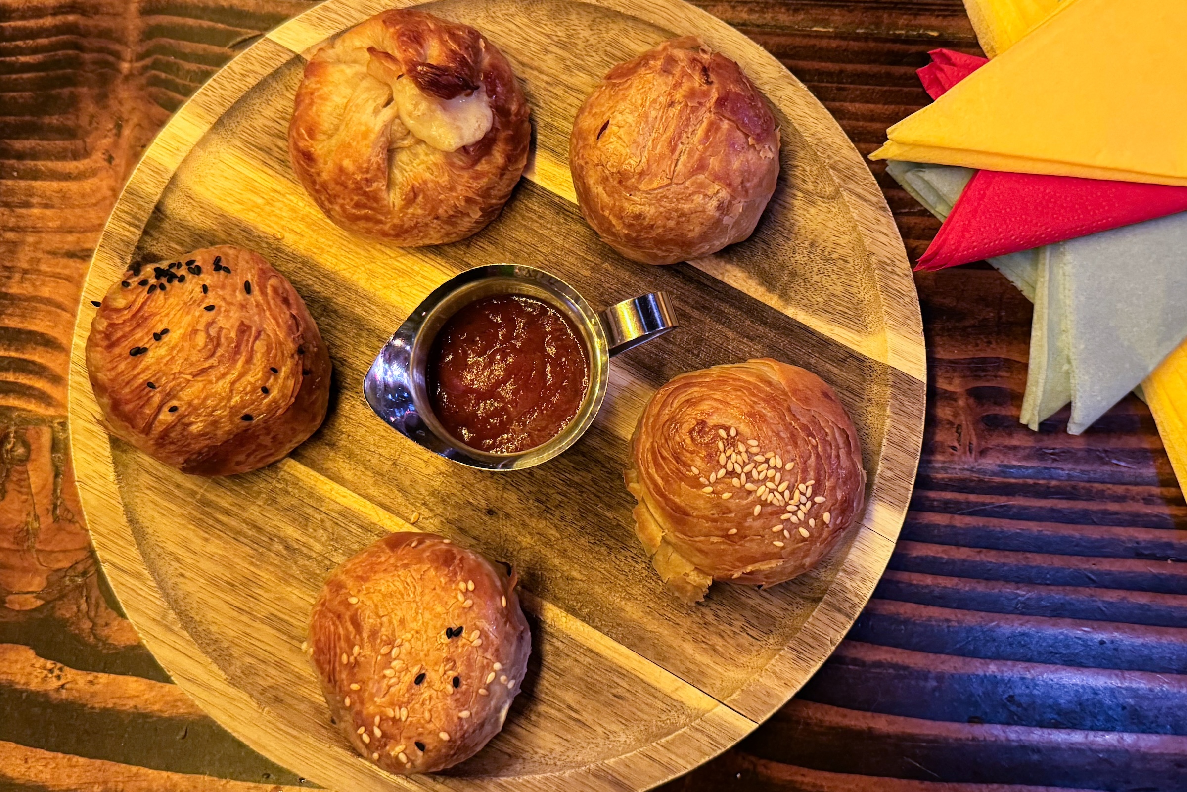 Five round, flaky Central Asian samsas (savory pastries) arranged on a circular wooden platter. Some are topped with black and white sesame seeds. A small metal pitcher of red tomato-based sauce sits in the center. Vibrant yellow and red napkins are visible in the upper right corner.