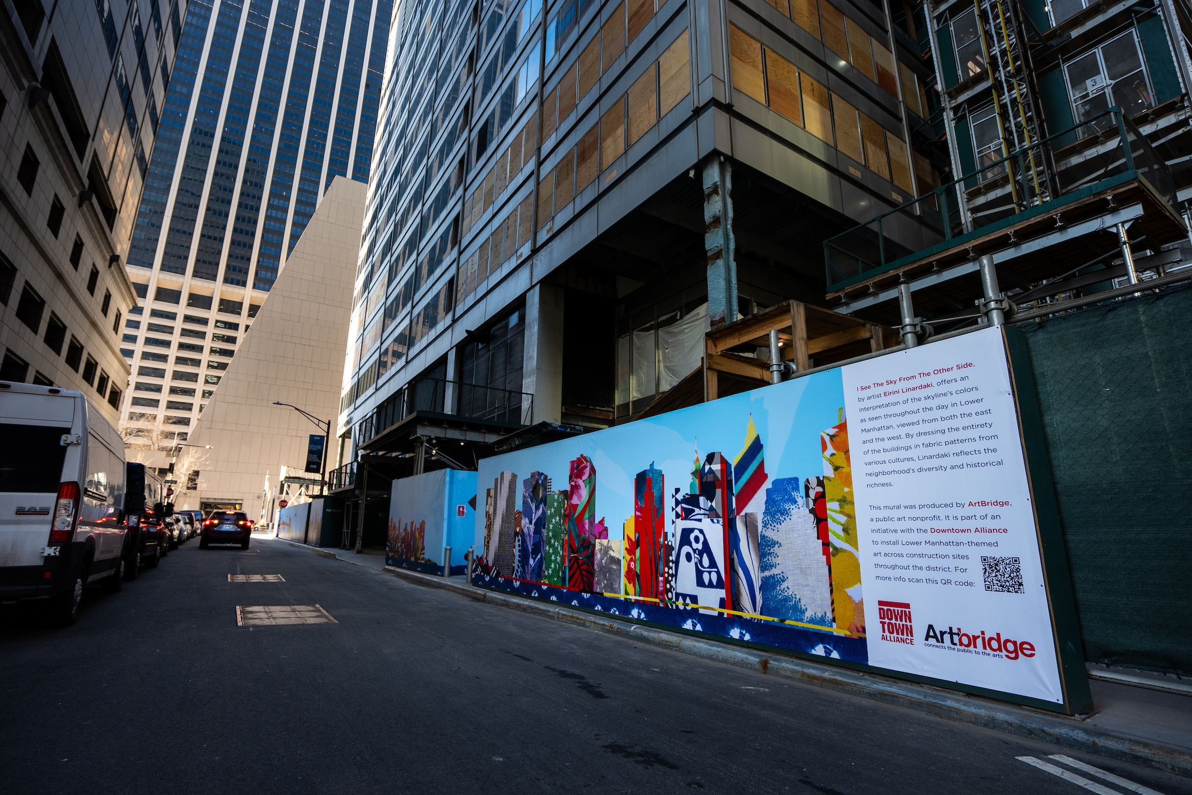 A wide shot looking down a narrow city street lined with tall buildings and parked vans. On the right, a long construction fence displays a colorful mural of a skyline made from diverse fabric patterns. The end of the mural features a white information panel with red and black text detailing the project, "I See The Sky From The Other Side" by Eirini Linardaki, and logos for ArtBridge and the Downtown Alliance. The street is in shadow, while sunlight hits the upper floors of the surrounding skyscrapers.