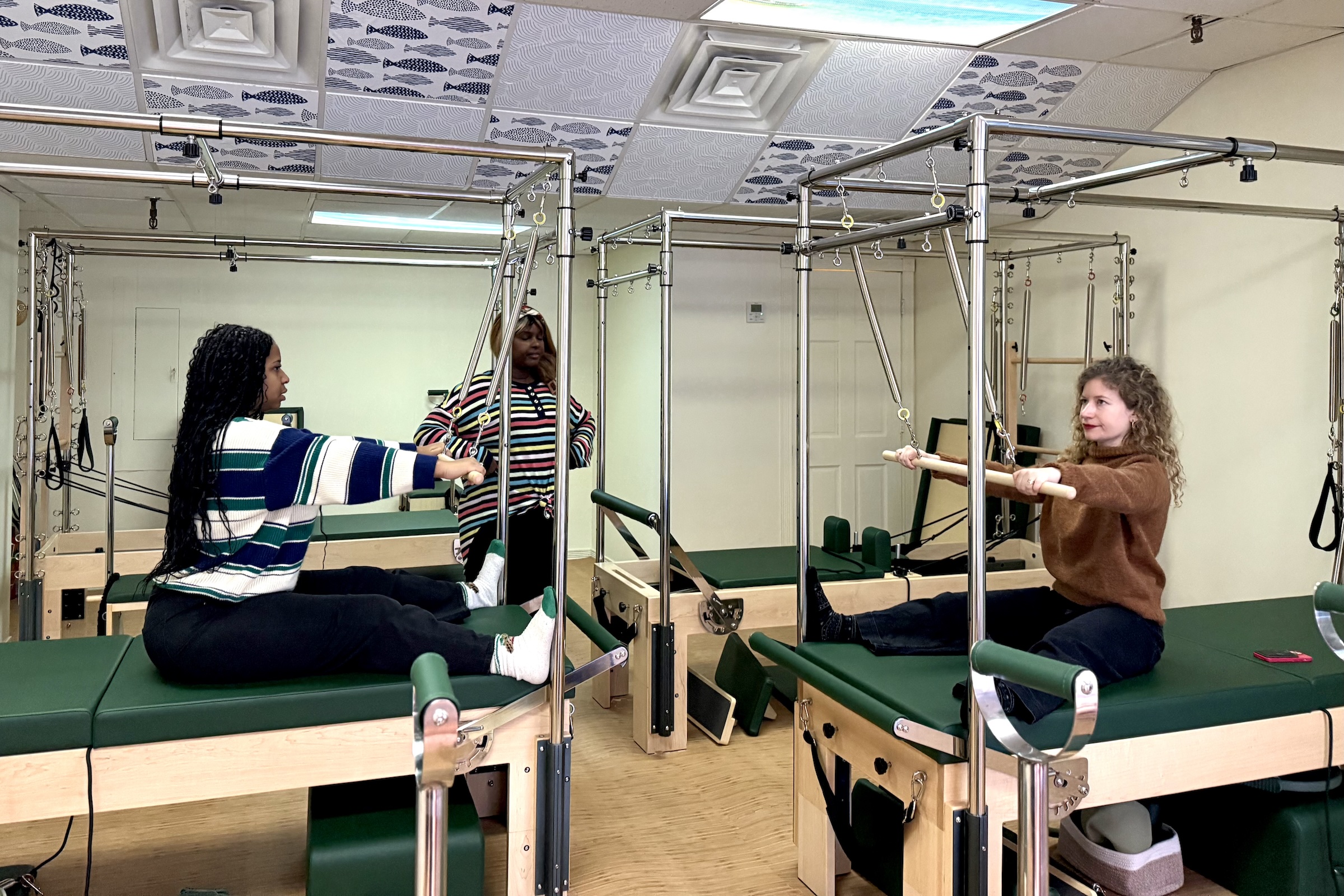 Three women are in a brightly lit Pilates studio, practicing on Cadillac reformers. In the foreground, two women sit facing each other on separate green upholstered machines, each holding a wooden push-through bar with their arms extended. One woman wears a green, white, and blue striped sweater, while the other wears a brown turtleneck. In the background, a third woman in a colorful striped shirt stands between the equipment, observing. The room features light wood floors and a unique ceiling tiled with a blue fish pattern.