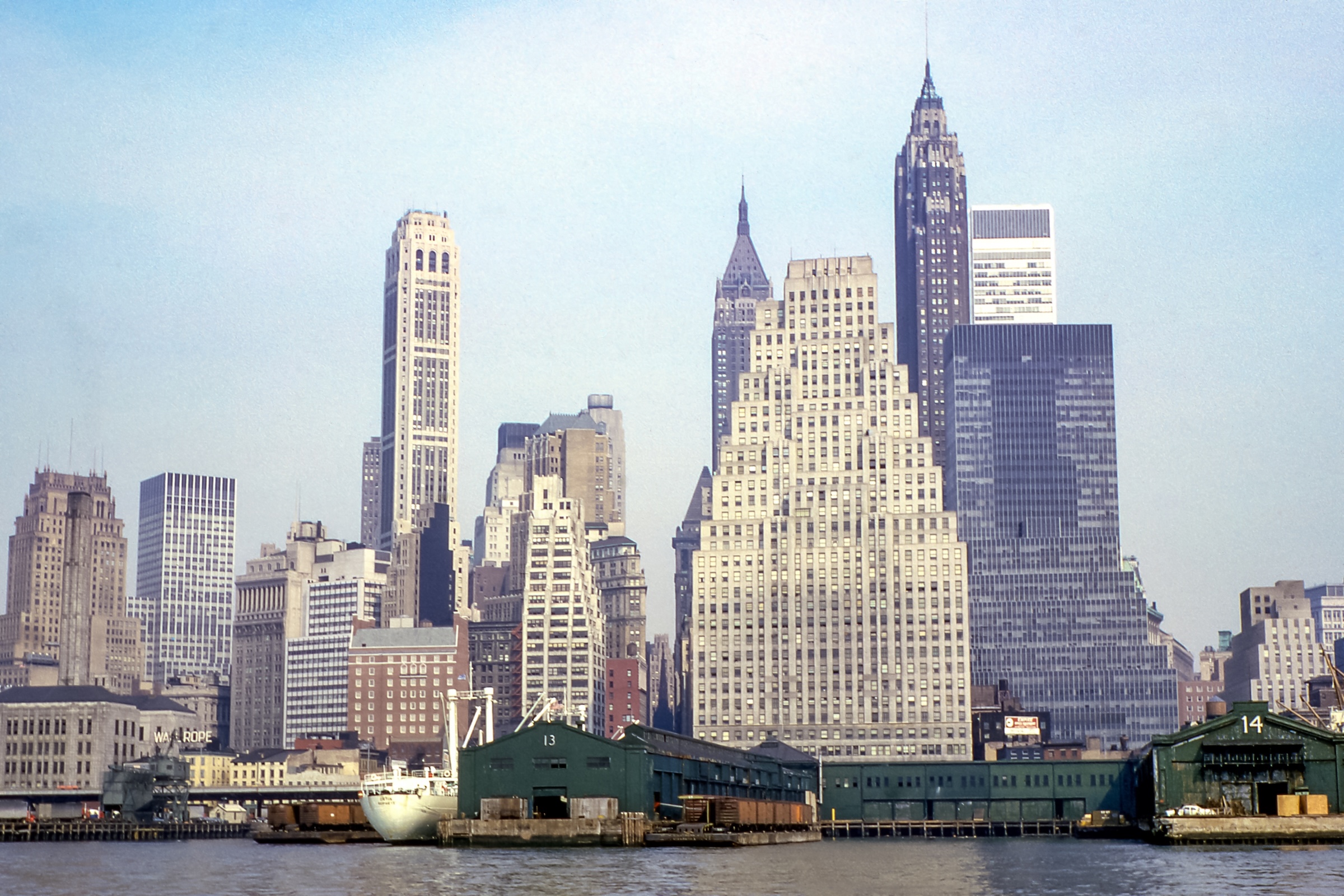 A wide landscape view of the Lower Manhattan skyline as seen from the water. The prominent, wedding-cake-style 120 Wall Street building stands in the center with its numerous tiered setbacks. In the background, other historic skyscrapers like 70 Pine Street and 20 Exchange Place pierce the horizon. In the foreground, green industrial piers and a white ship sit along the East River.