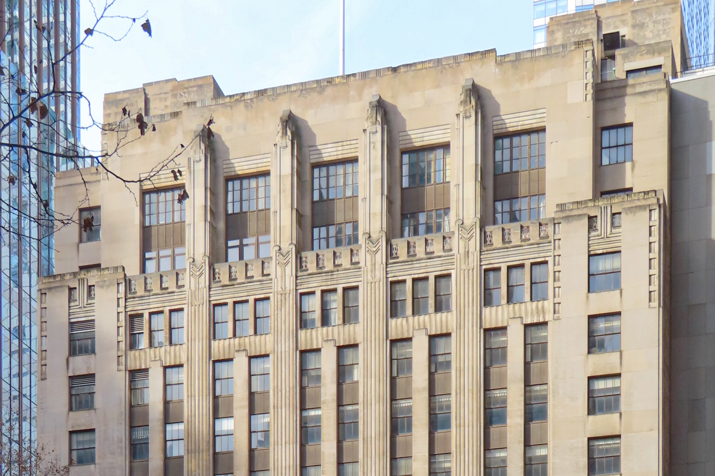 A view of the upper floors of a limestone Art Deco building. The architecture emphasizes verticality with prominent, fluted pilasters that extend past the roofline. Below the top windows, there is a decorative stone frieze featuring stylized, geometric floral or leaf motifs typical of the period.