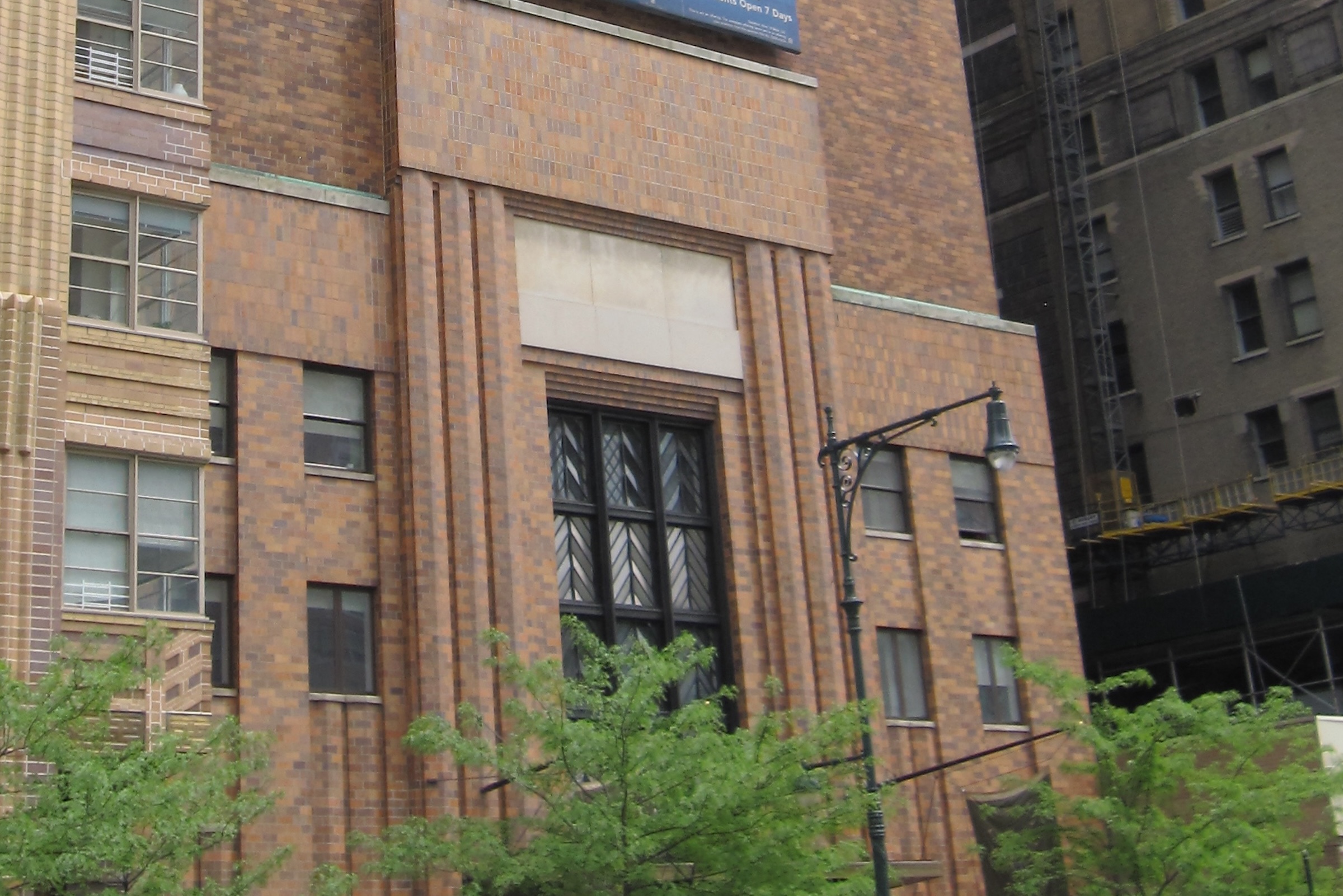 A medium shot of a section of a brown and tan brick Art Deco building. The facade features vertical recessed channels and a large, multi-paned window with a distinct chevron pattern in the metalwork. Green tree branches are visible in the foreground, partially obscuring the lower levels.