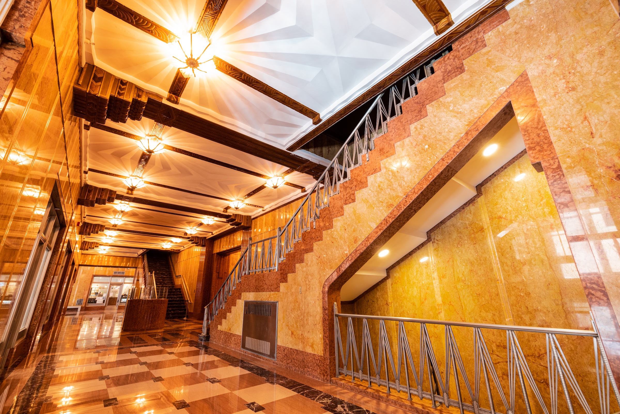 A wide-angle interior shot of a grand Art Deco lobby. The space is defined by polished marble walls in shades of amber and rose, a patterned stone floor, and a dramatic staircase with a geometric metal railing. The ceiling features ornate, stepped coffers with glowing, starburst-style light fixtures.