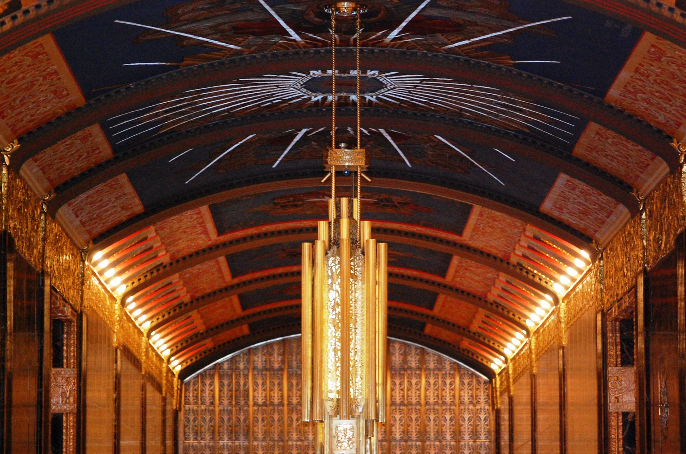 A low-angle shot looking up at a magnificent, barrel-vaulted ceiling in a grand hallway. The ceiling is painted a deep midnight blue and adorned with gold leaf sunbursts and intricate murals. A massive, tiered gold chandelier with a geometric, pipe-like design hangs in the foreground, while the side walls are lined with rows of warm, glowing lights that emphasize the architectural curve.