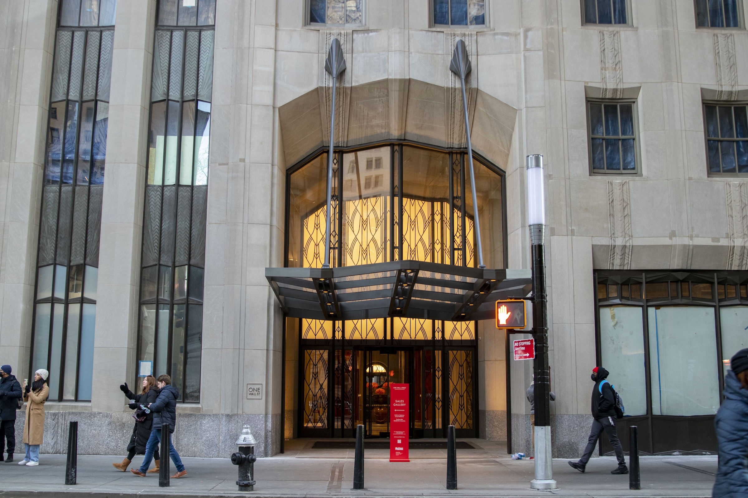 An eye-level view of a modern entrance to a classic limestone building. It features a dark metal canopy and a large glass entryway with a backlit, intricate gold screen in a repeating geometric "Red Room" pattern. Flanking the entrance are tall windows with distinctive rippled or "curtain" glass typical of Ralph Walker's design.