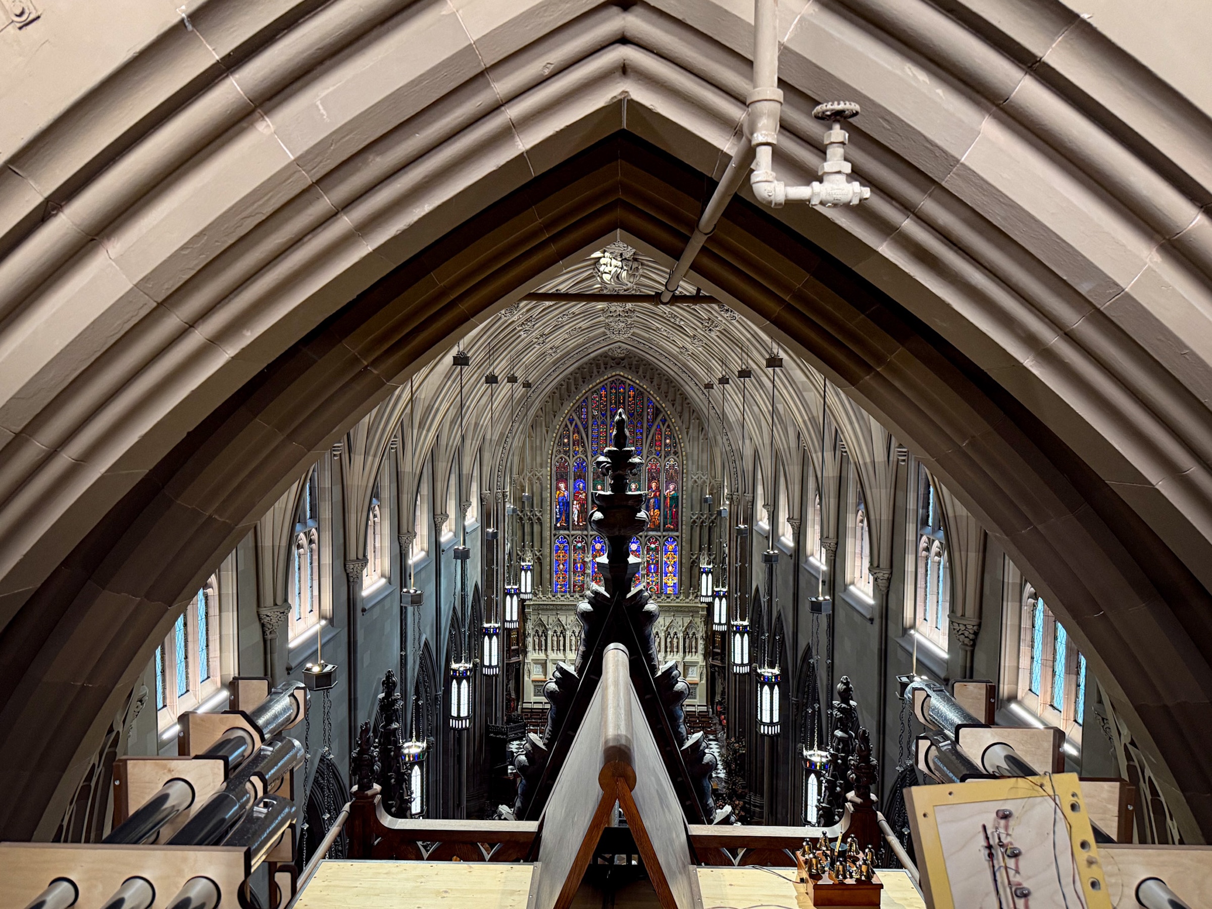 An elevated, "bird’s-eye" perspective looking down the long nave of the cathedral from the organ loft. Ornate stone arches frame the view, leading the eye toward a large, colorful stained-glass window at the far end. In the foreground, the tops of organ pipes and mechanical components are visible, emphasizing the height and scale of the building.