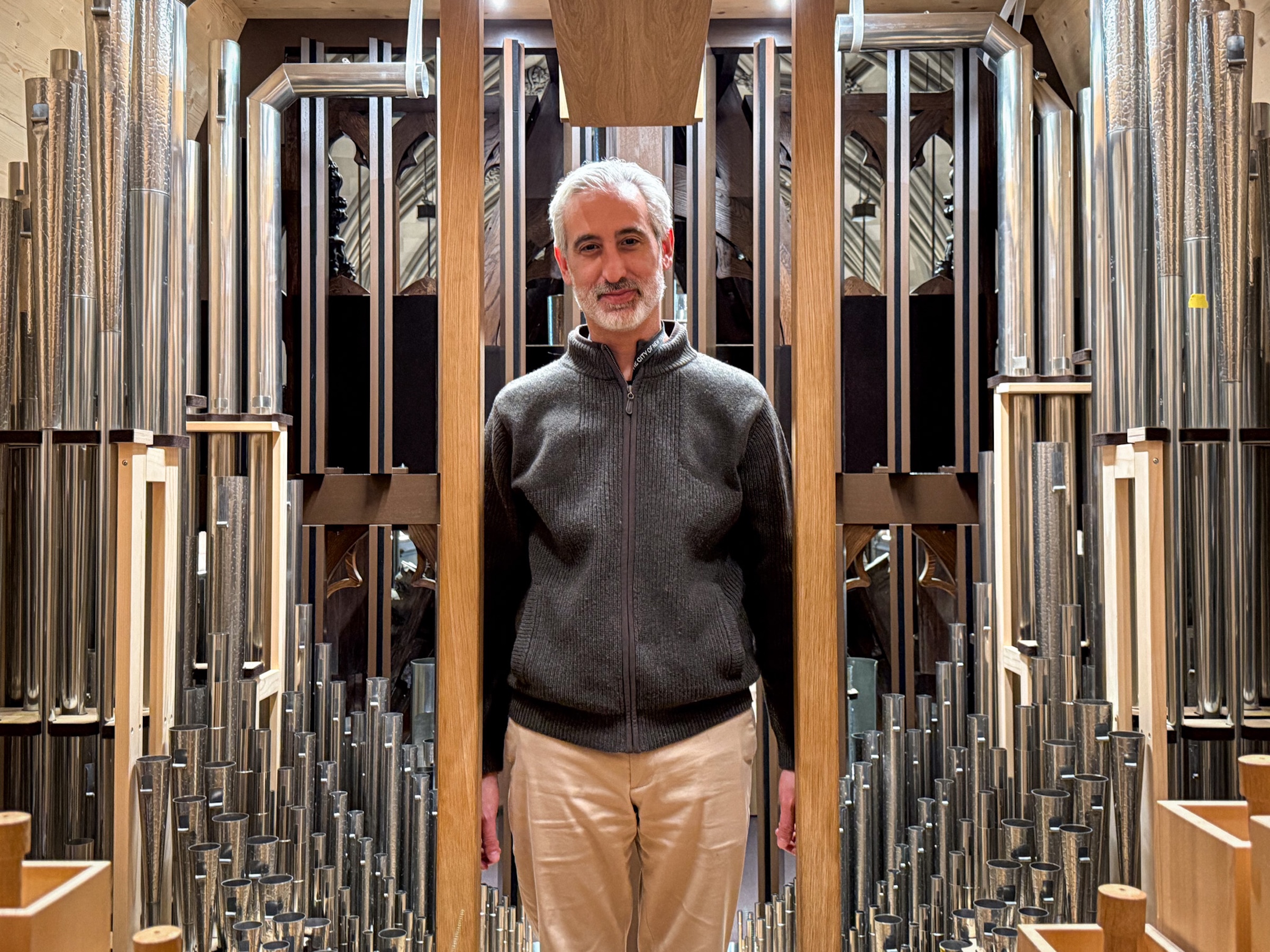 A portrait of the same man standing amidst the internal workings of the organ. He is flanked by hundreds of vertical metal pipes of varying sizes, ranging from small tubes at his waist to large, thick pipes extending above his head. The wooden framework and the complex arrangement of the pipes create a dense, industrial aesthetic within the sacred space.
