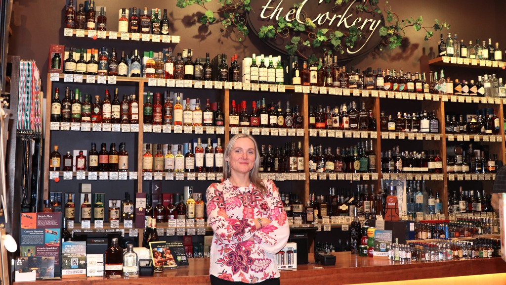 A woman with blonde hair and a floral blouse stands with her arms crossed in front of a massive, floor-to-ceiling wooden shelving unit. The shelves are densely packed with hundreds of bottles of wine and spirits. Above her, a decorative sign reads "the Corkery" surrounded by faux grapevines.