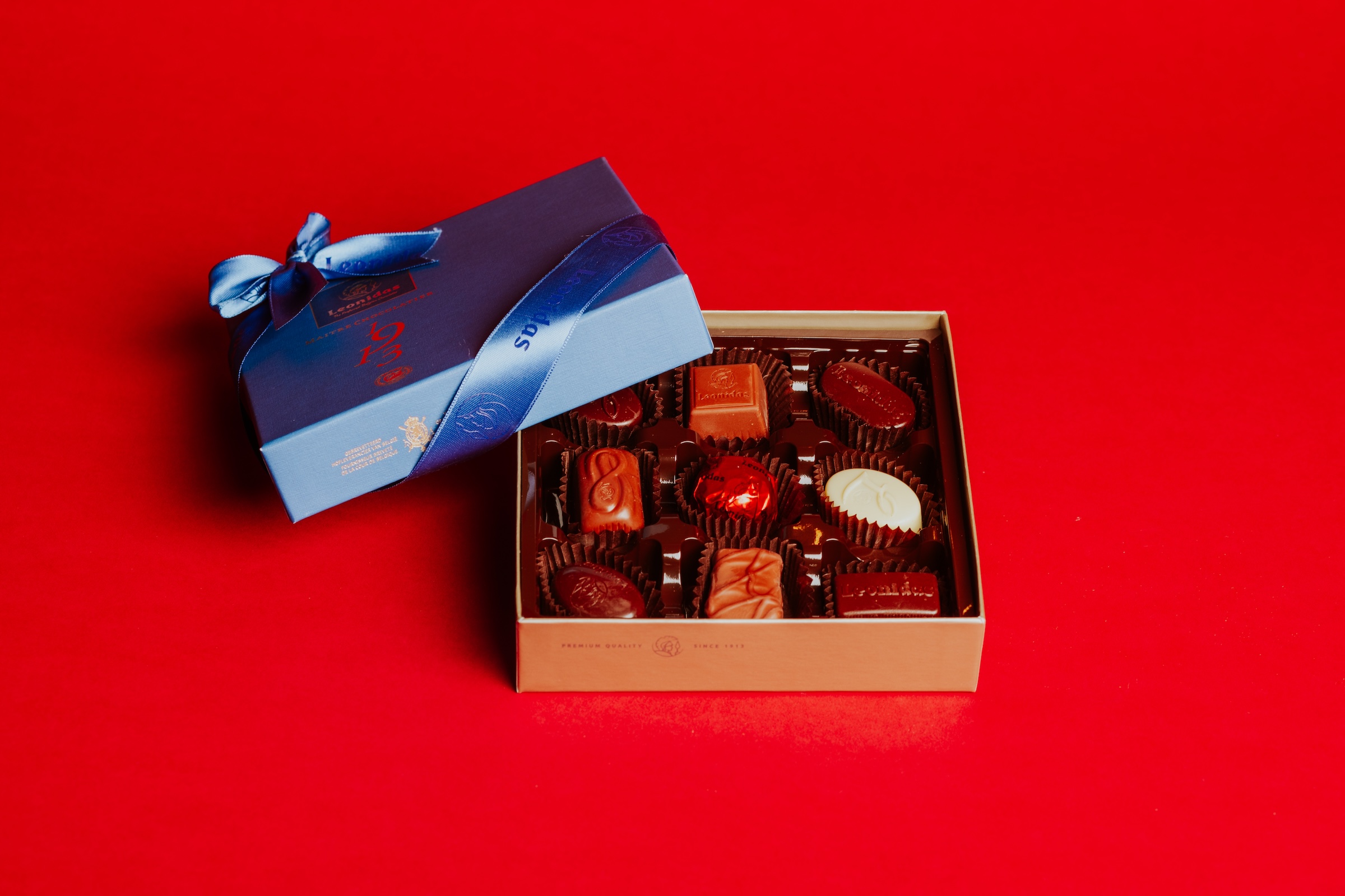 A square, gold-colored box of Leonidas Belgian chocolates sits open on a bright red surface. The box contains an assortment of dark, milk, and white pralines in paper cups. A blue lid with a matching satin ribbon and bow leans against the side of the box.
