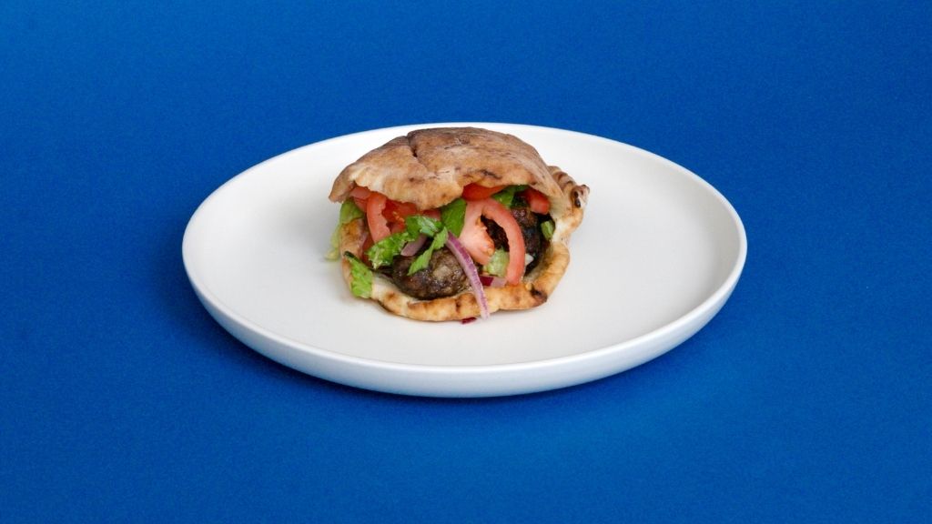 A single pita sandwich filled with grilled meat, sliced tomatoes, red onions, and fresh greens sits centered on a plain white plate. The entire composition is set against a solid, vibrant blue background.