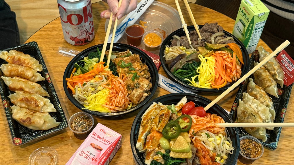A top-down view of a wooden table covered in a vibrant Korean feast. There are three large bowls of bibimbap filled with colorful ingredients like kimchi, spinach, bean sprouts, and protein, alongside two trays of golden-brown pan-fried mandu (dumplings). The spread is rounded out by a can of Diet Coke, small cartons of strawberry and melon milk, and various dipping sauces.