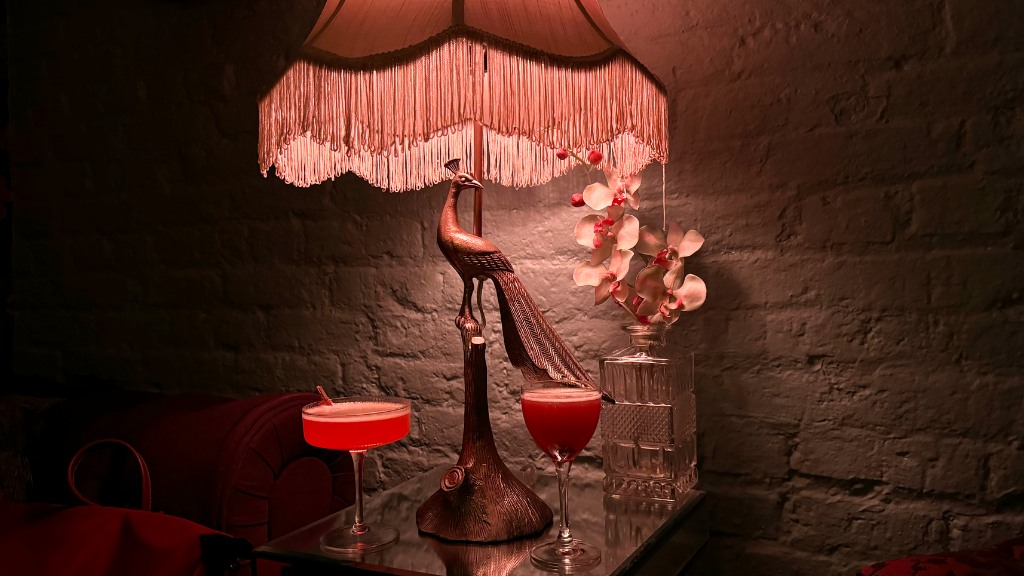 A moody, dimly lit scene featuring two red cocktails on a reflective side table, anchored by an ornate peacock lamp against a white brick wall.