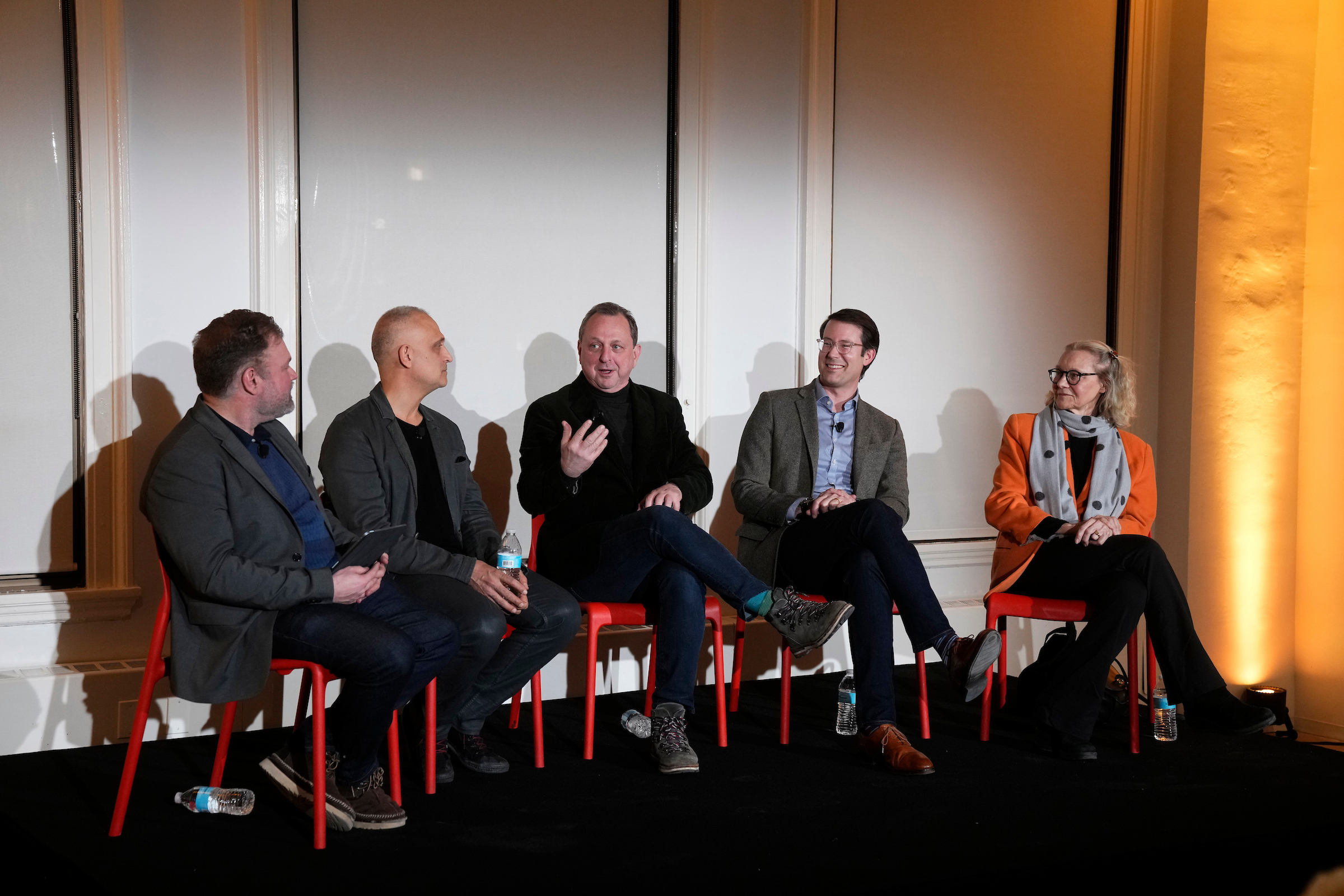 A panel of five people sits on modern red chairs on a low black stage during an evening event titled "From Cathedral of Commerce to Catalyst for Culture." From left to right: Justin Rivers (moderator), Ron Castellano, Gregg Pasquarelli (speaking), Nate Rogers, and Carol Willis. They are dressed in business-casual attire, including blazers and sweaters. The background consists of white paneled walls illuminated by warm, amber architectural lighting.