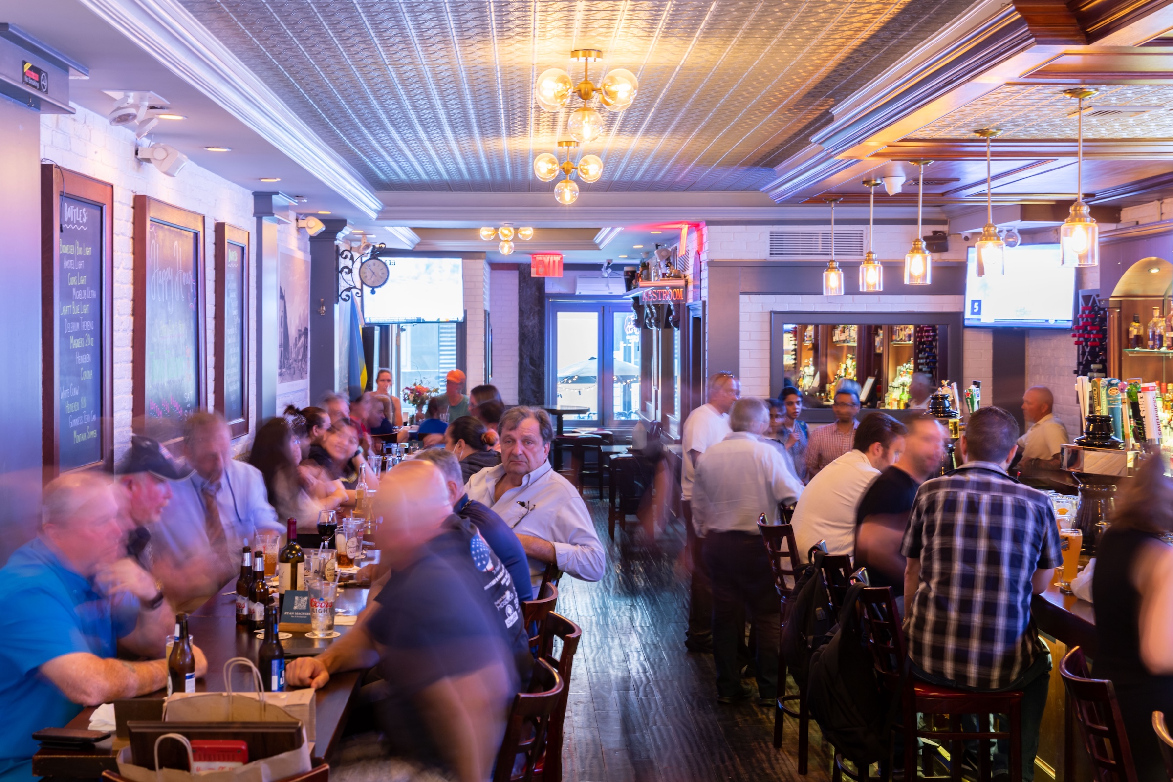 A bustling, brightly lit long-bar and restaurant interior with a warm atmosphere. Patrons are seated along a dark wood bar on the left and a series of high-top stools on the right. The ceiling features ornate metallic tiles and glowing globe chandeliers. On the left wall, several chalkboard menus are mounted against white brick. The long, narrow space creates a deep perspective leading toward a windowed entrance in the back.